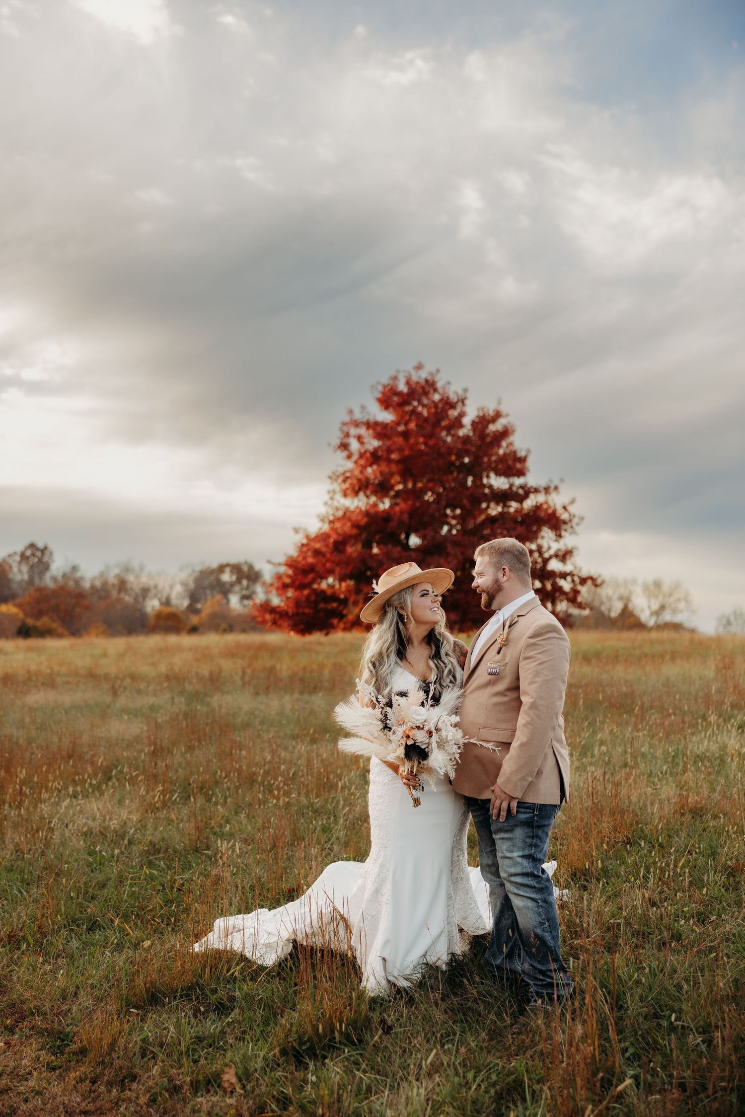 Couple in boho wedding attire embracing in autumn field with red tree backdrop under dramatic cloudy sky.