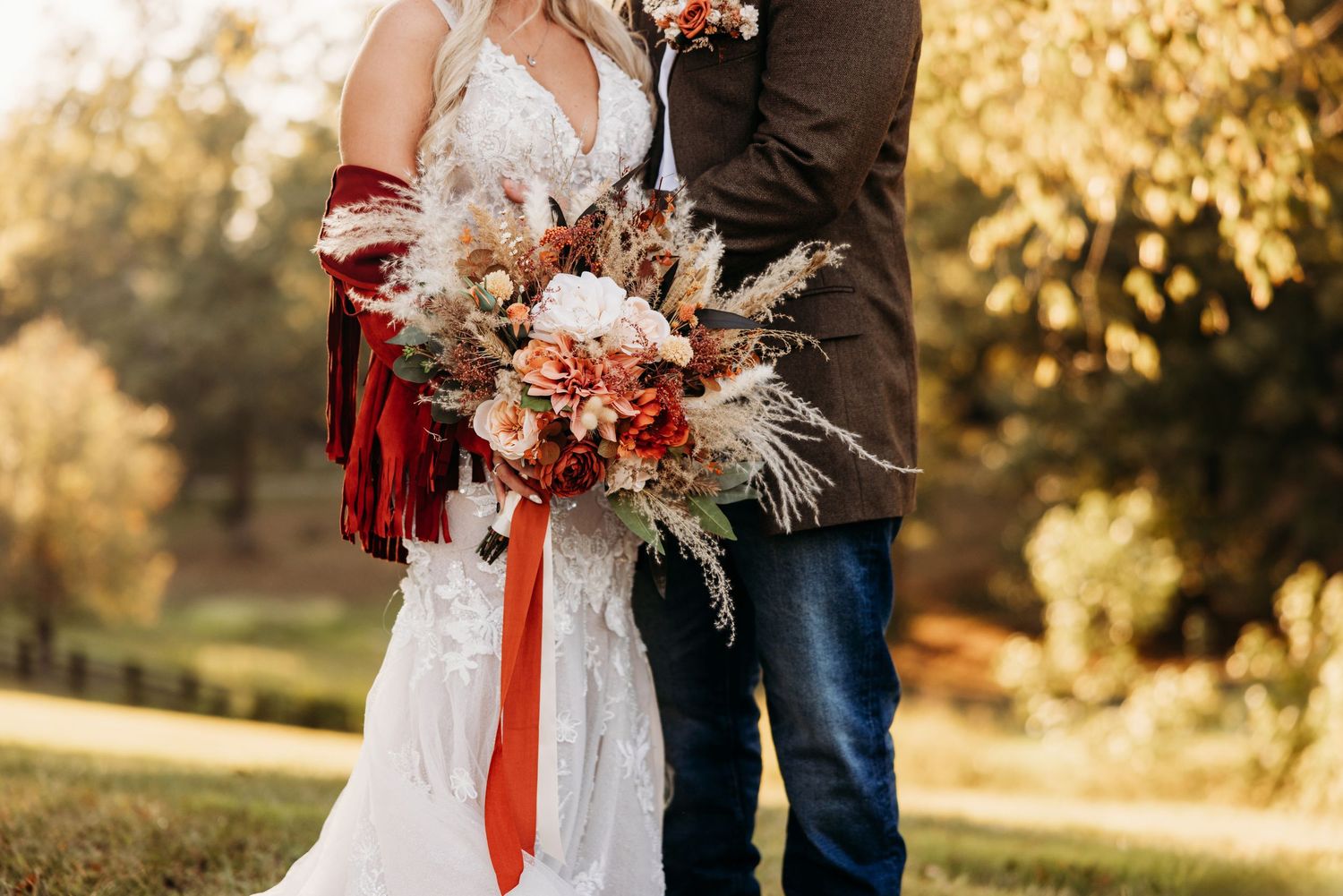 A couple stands together outdoors during golden hour holding a bouquet with red and white flowers and dried pampas grass.