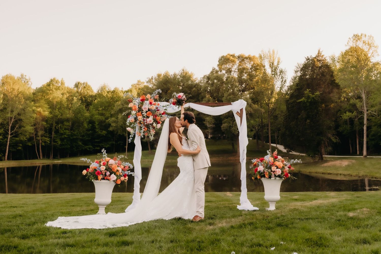 Couple kissing under a floral wedding arch with colorful blooms and white draping, set on a green lawn near a pond.