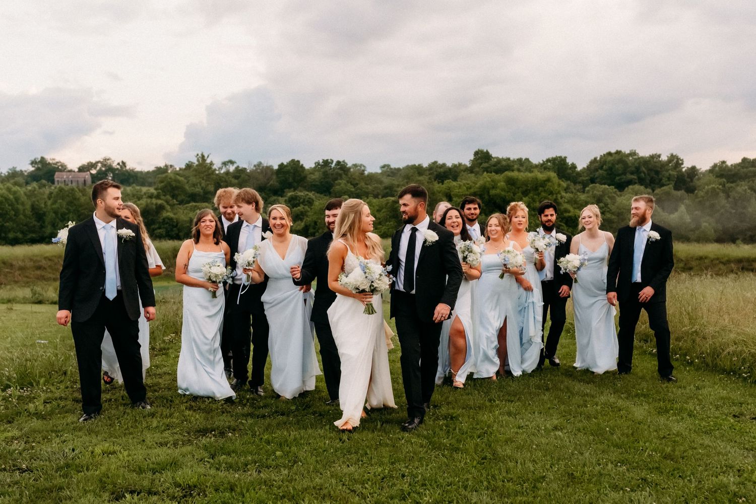 Wedding party walks outdoors in formal attire, bridesmaids in white gowns and groomsmen in black suits, on a green lawn.