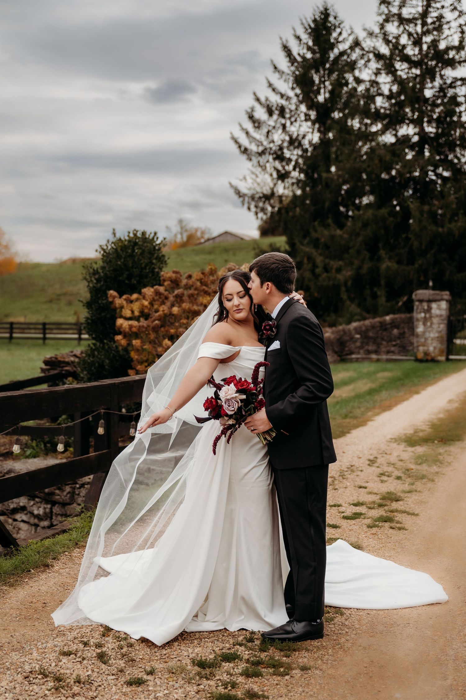 Bride in off-shoulder white gown with veil and groom in black tuxedo sharing romantic moment on gravel path outdoors.
