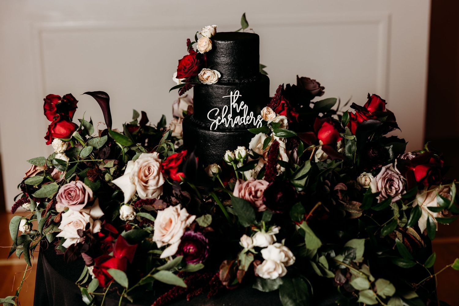 Black three-tier wedding cake with white calligraphy script, surrounded by lush red and blush roses and dark greenery.