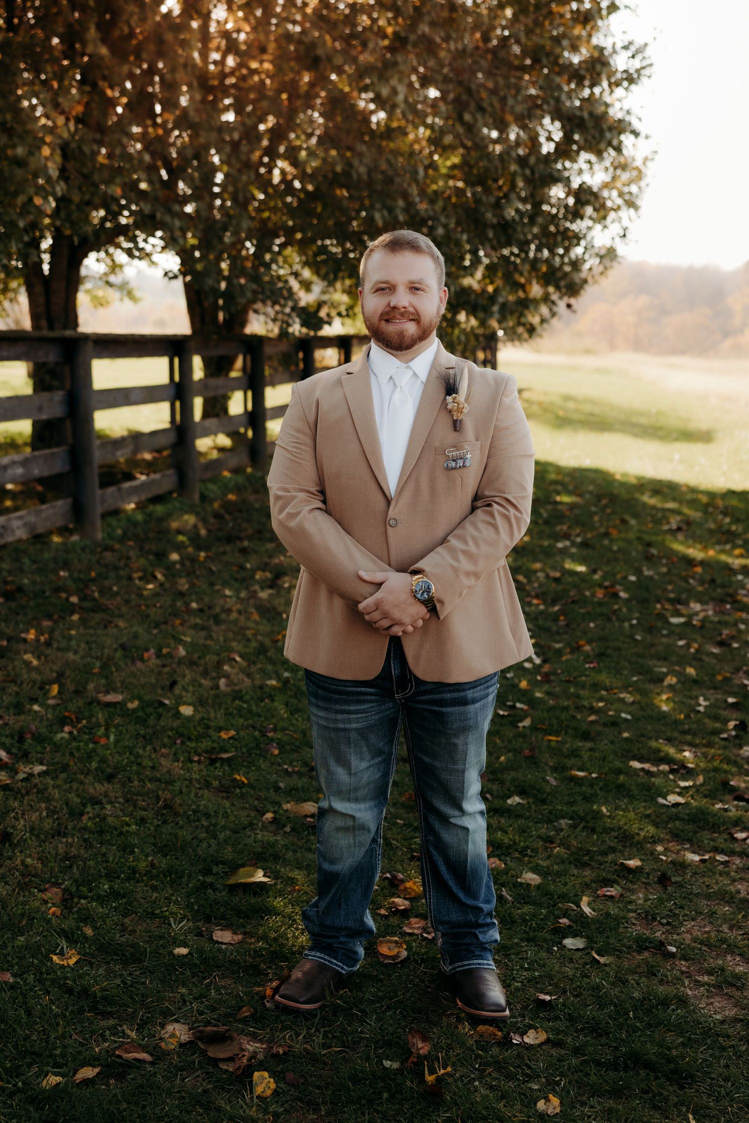 Groom in tan blazer and jeans with boutonniere stands smiling outdoors at autumn farm wedding venue.