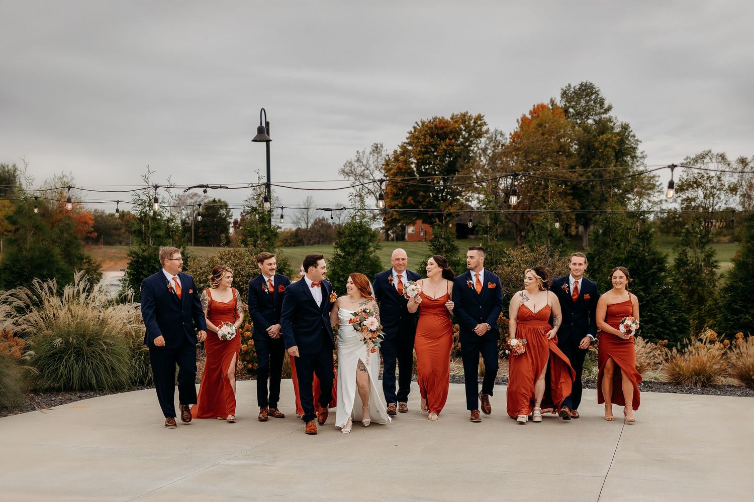 A wedding party in navy suits and rust-colored dresses poses outdoors.