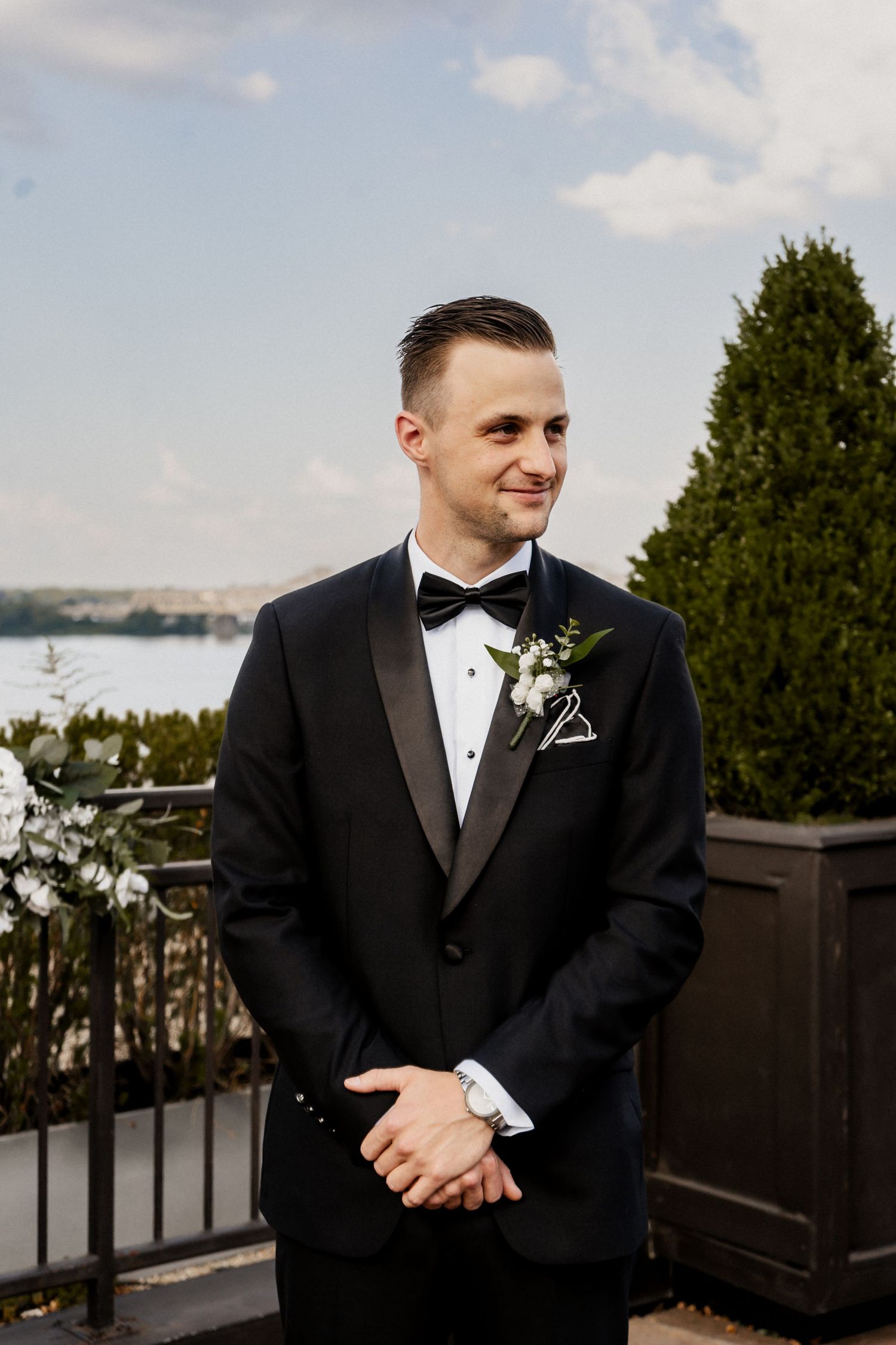 Groom in classic black tuxedo and bowtie smiles while standing on outdoor terrace with water view.