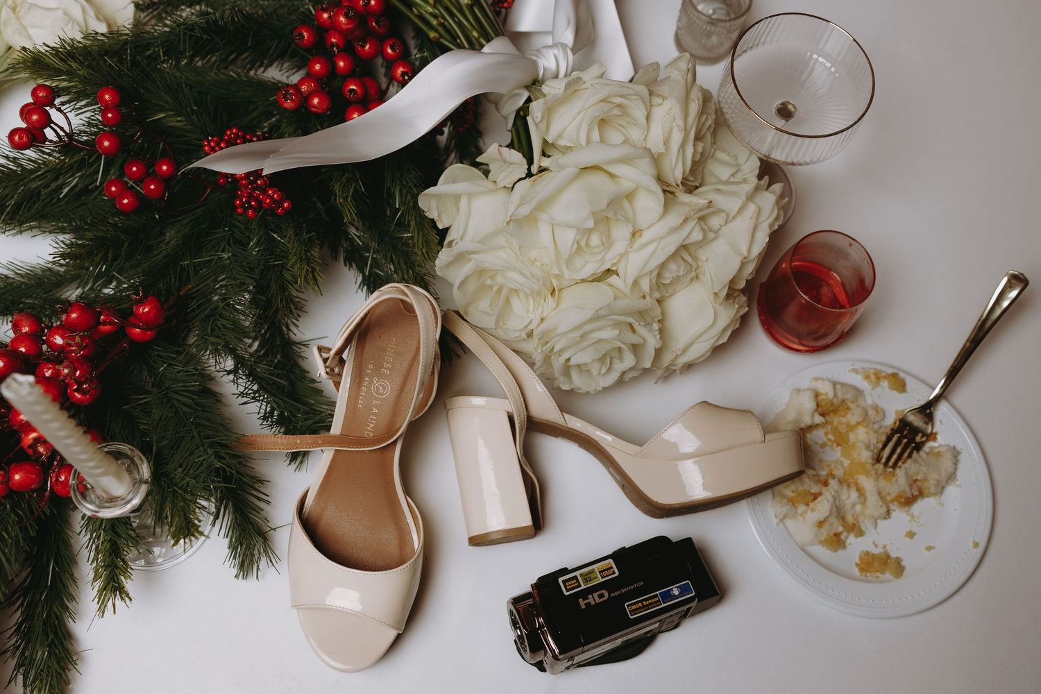 Wedding details flatlay with white shoes, evergreen branches, and red berries.