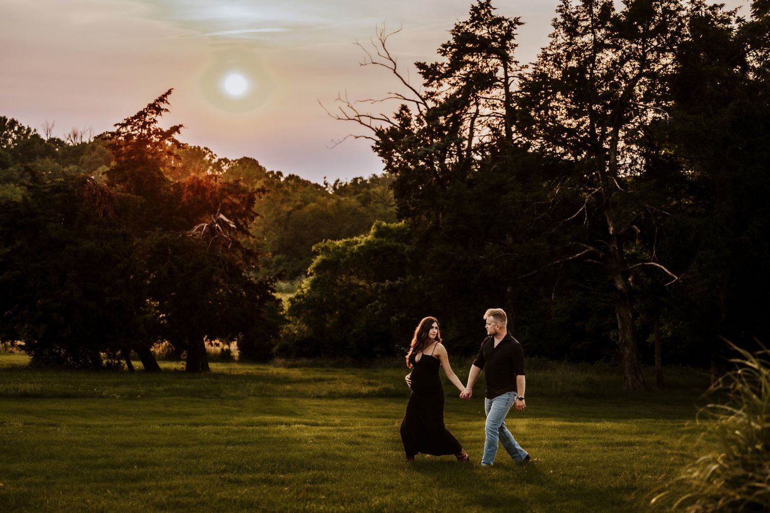 Silhouettes of a couple walking in a field at sunset with trees in the background.