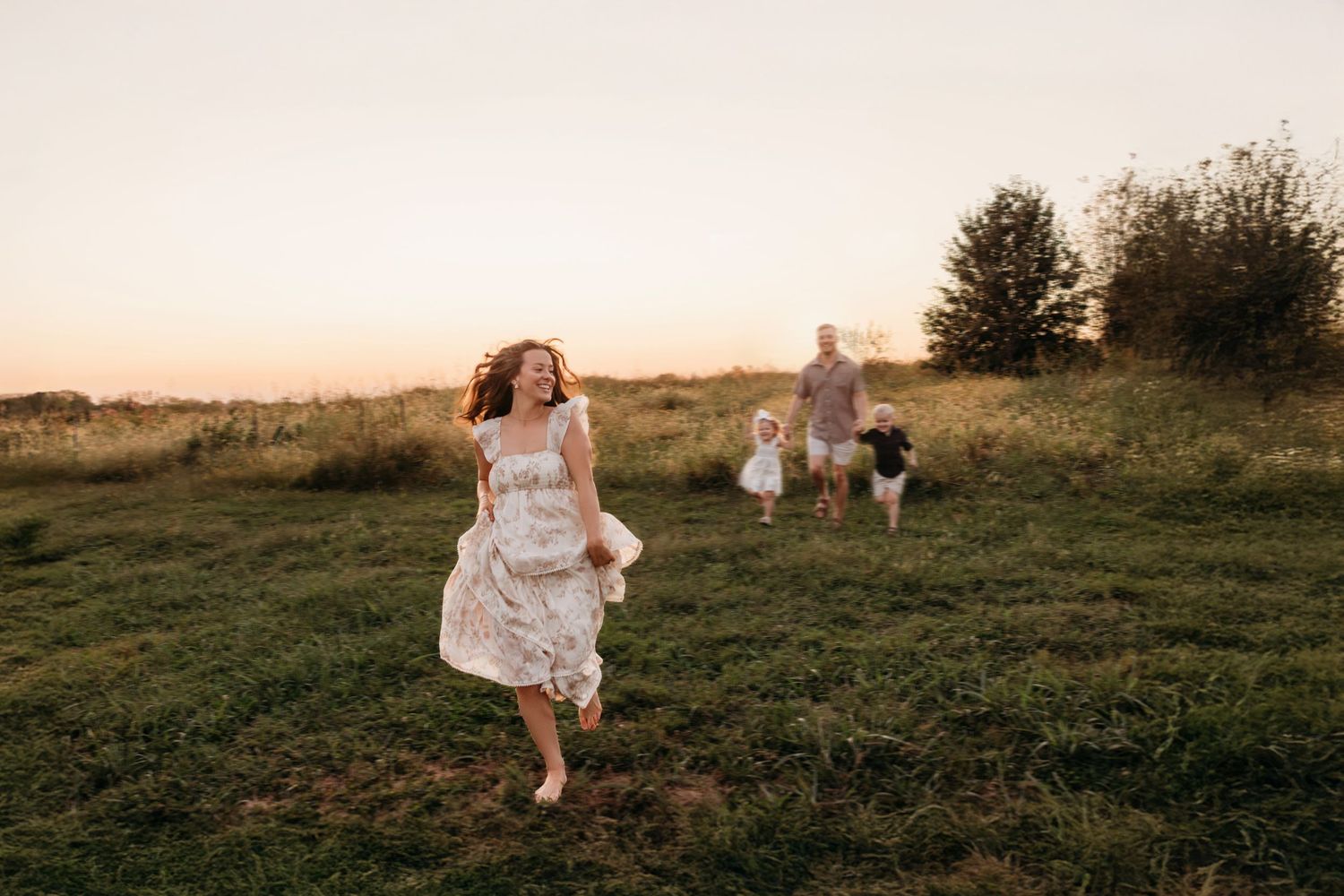 People in white dresses run through a grassy field at sunset creating a dreamy summer scene.