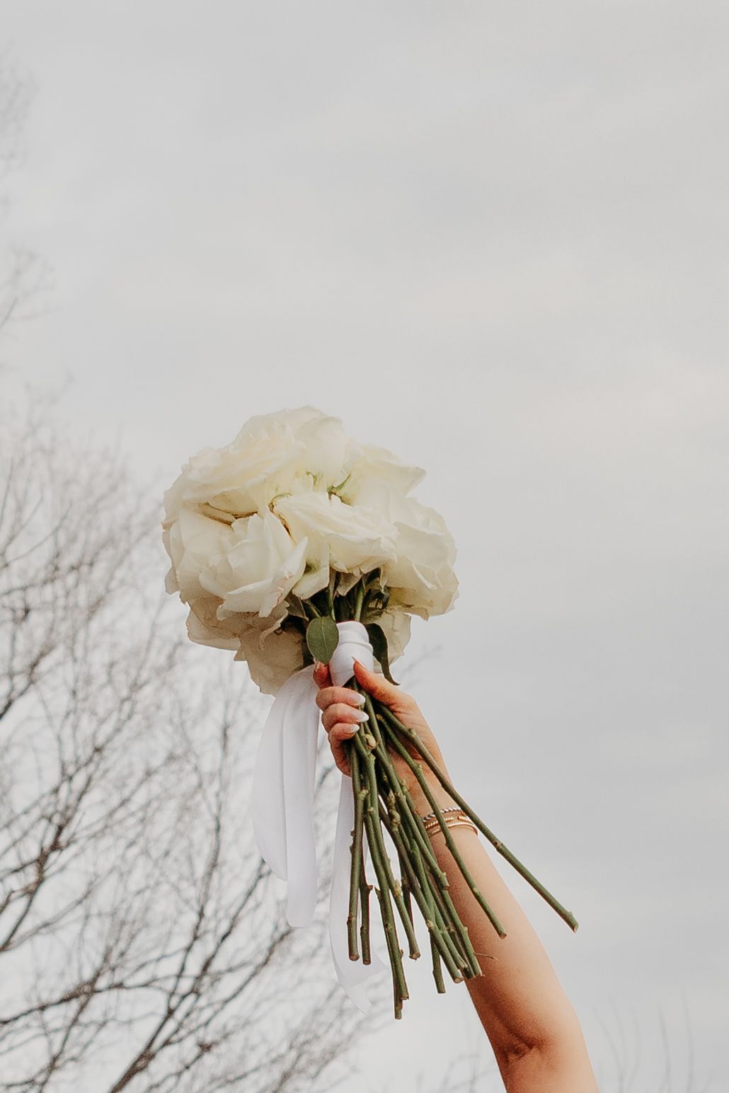 Hand holding a bridal bouquet of white roses with ribbon against a cloudy sky.