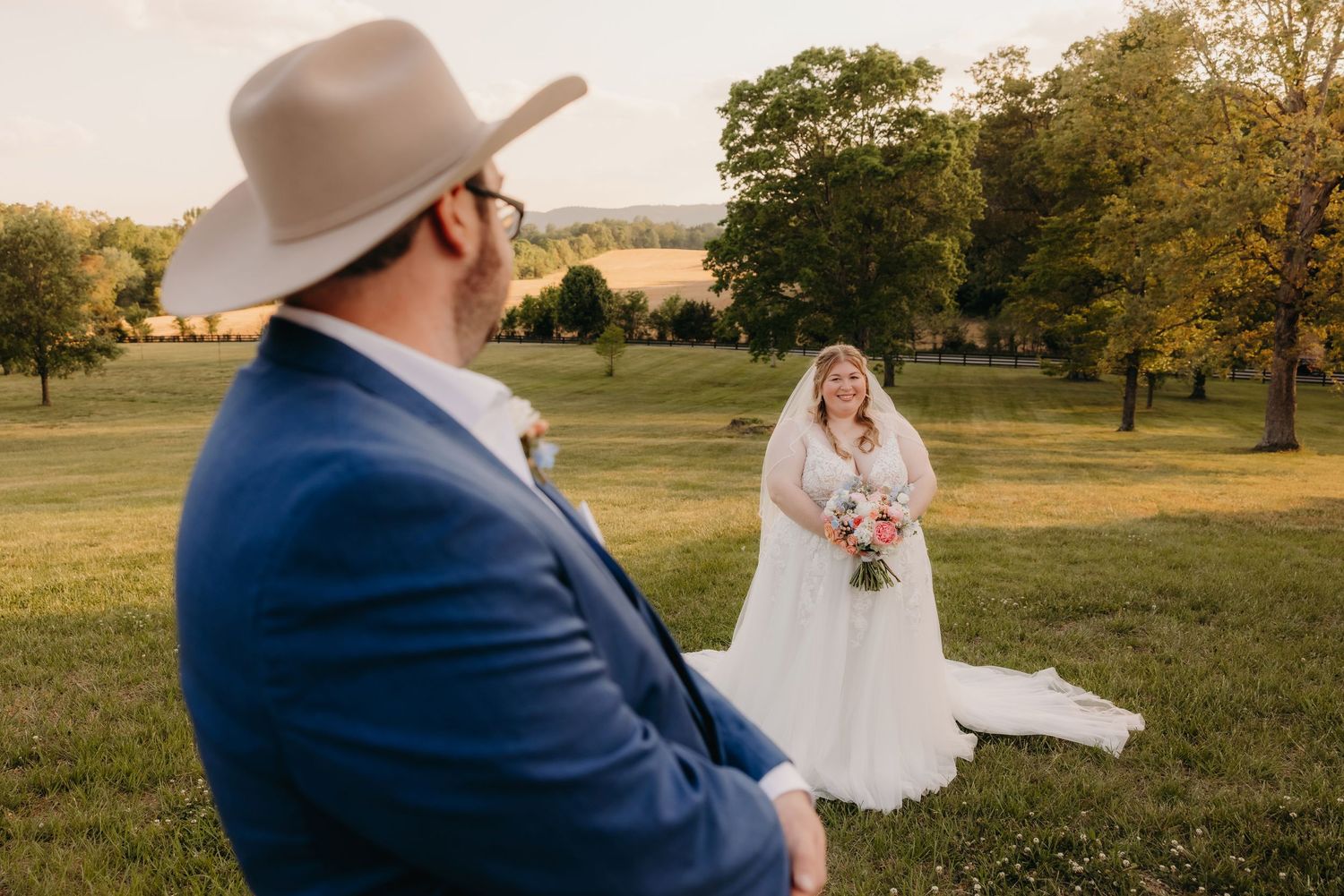 Groom in blue suit and cowboy hat watches bride in white gown standing in sunlit green field.