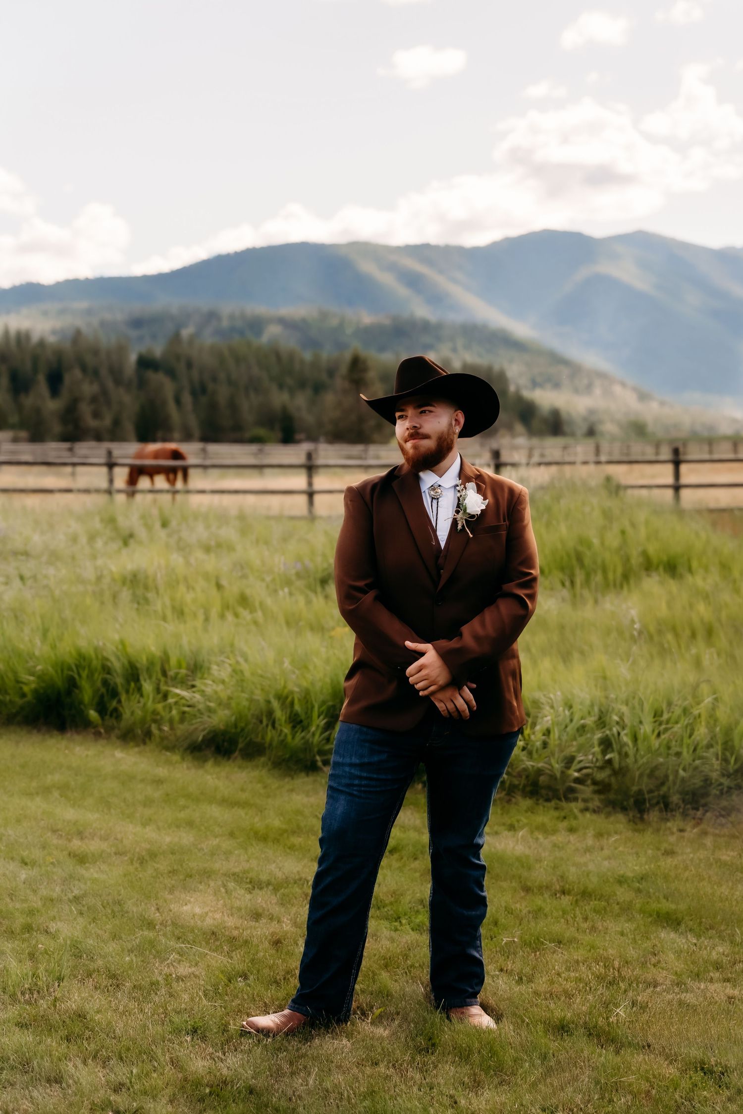 A groom in a brown blazer and cowboy hat stands in a scenic mountain ranch field with a horse.
