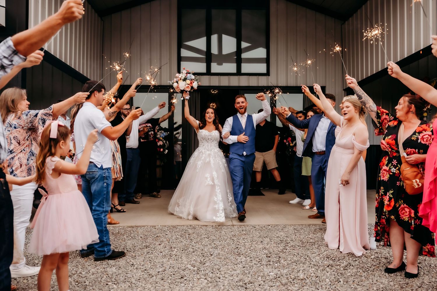 Bride and groom exit wedding venue through sparkler tunnel surrounded by cheering guests and wedding party.