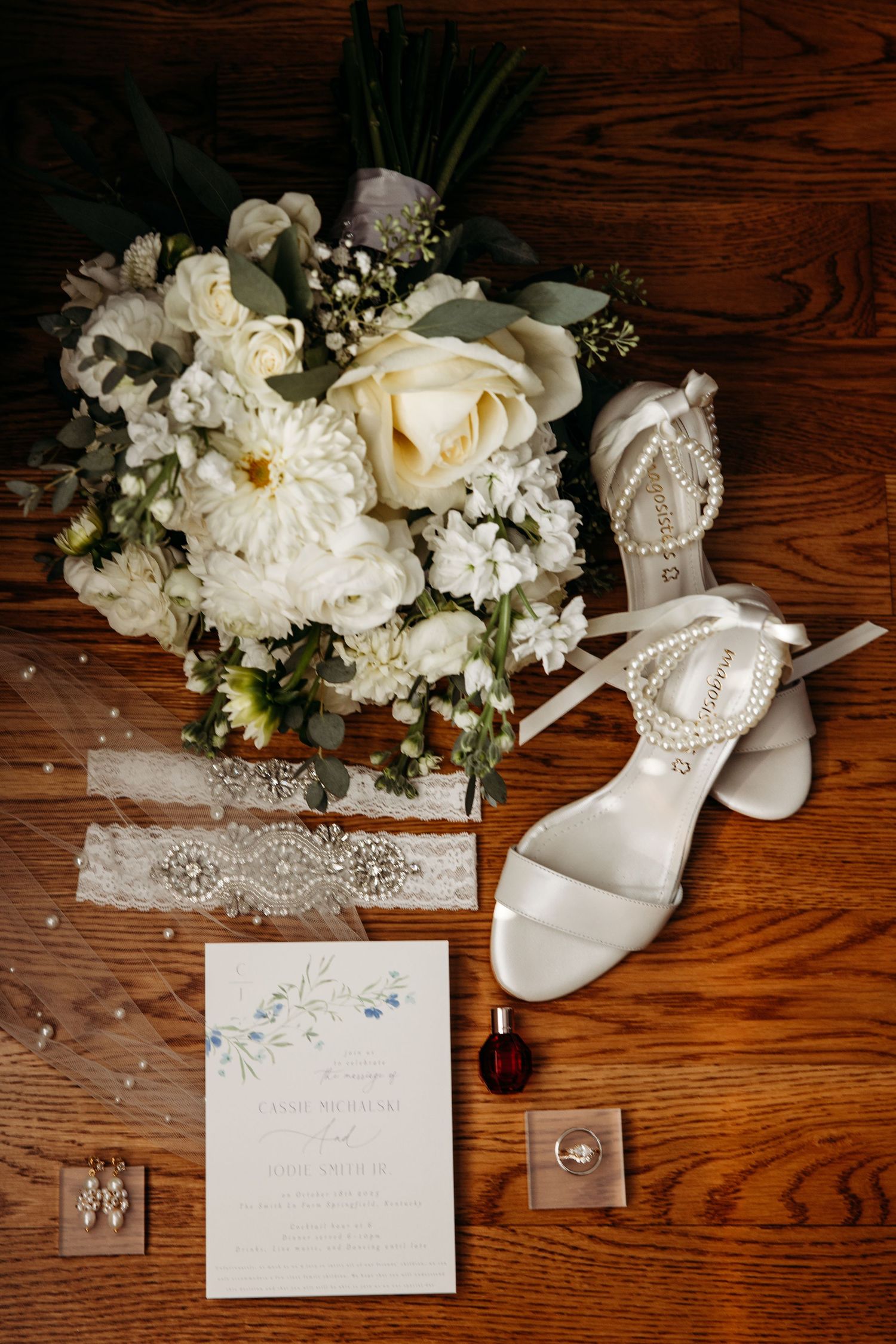Elegant bridal flat lay featuring white floral bouquet, pearl heels, lace garter, wedding invitation, earrings, and ring on wood floor.