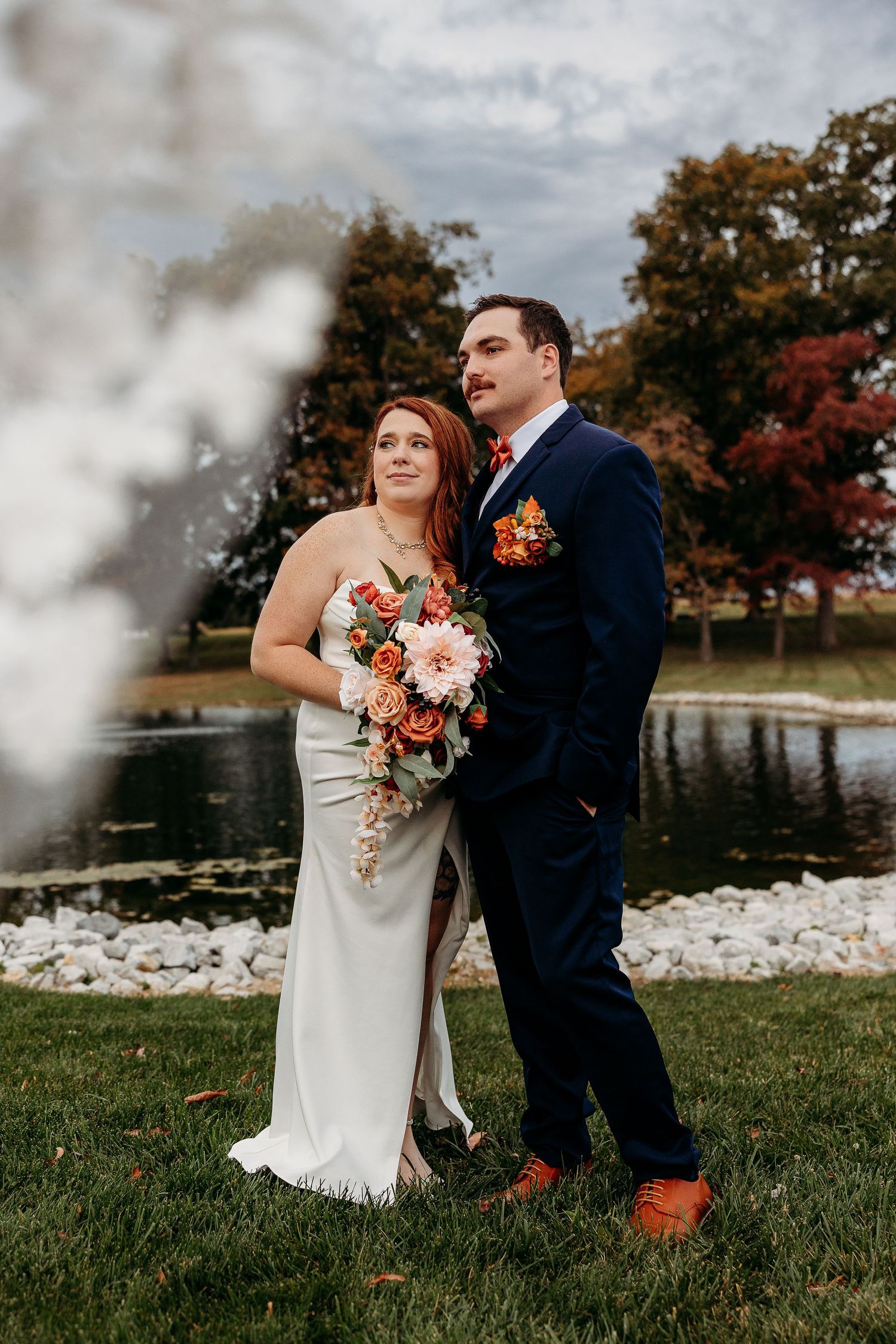 Bride in white gown holding orange floral bouquet stands with groom by pond amid autumn foliage.