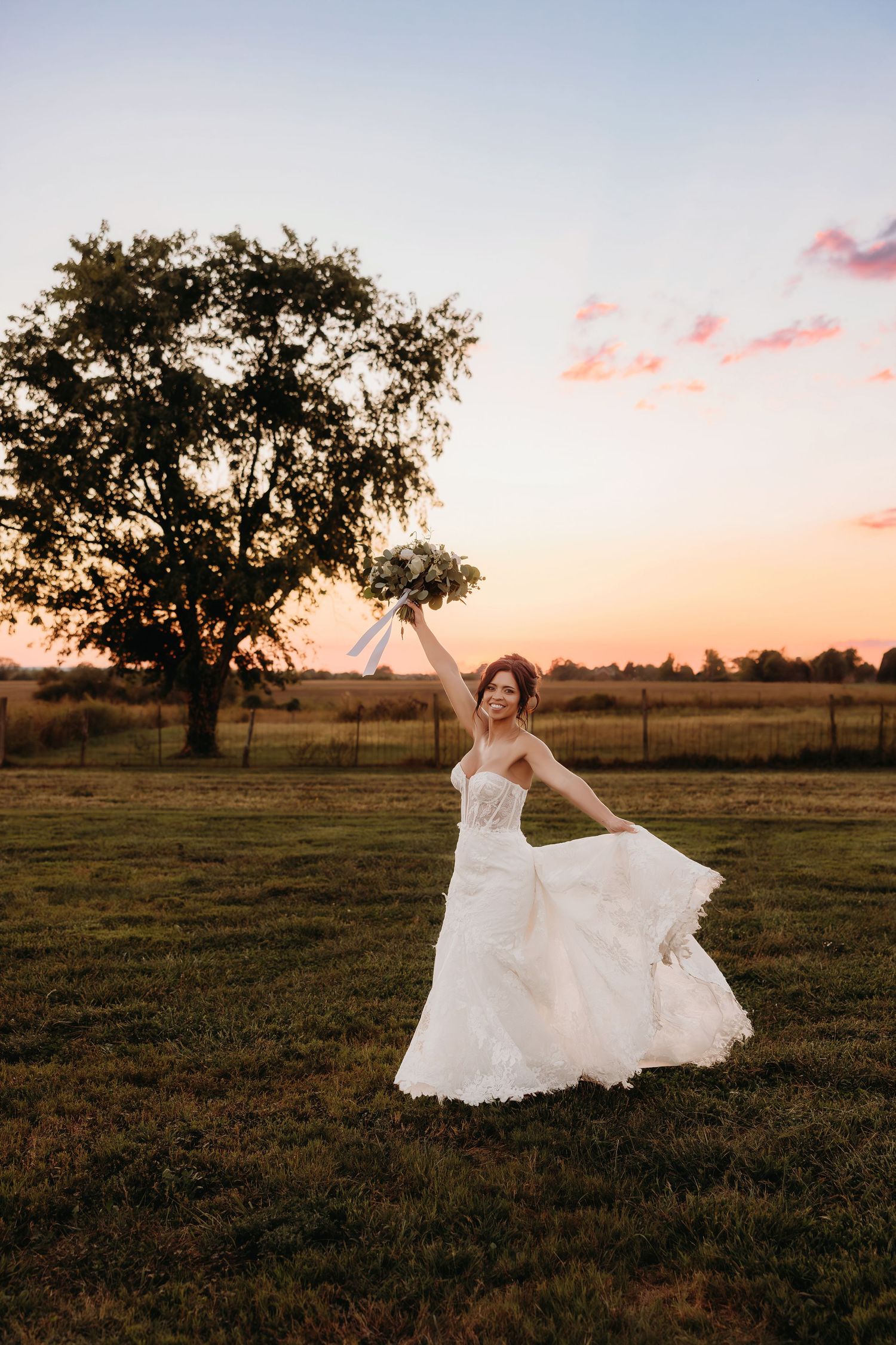 Joyful bride in white lace gown twirling at sunset in a green field with a large tree backdrop.