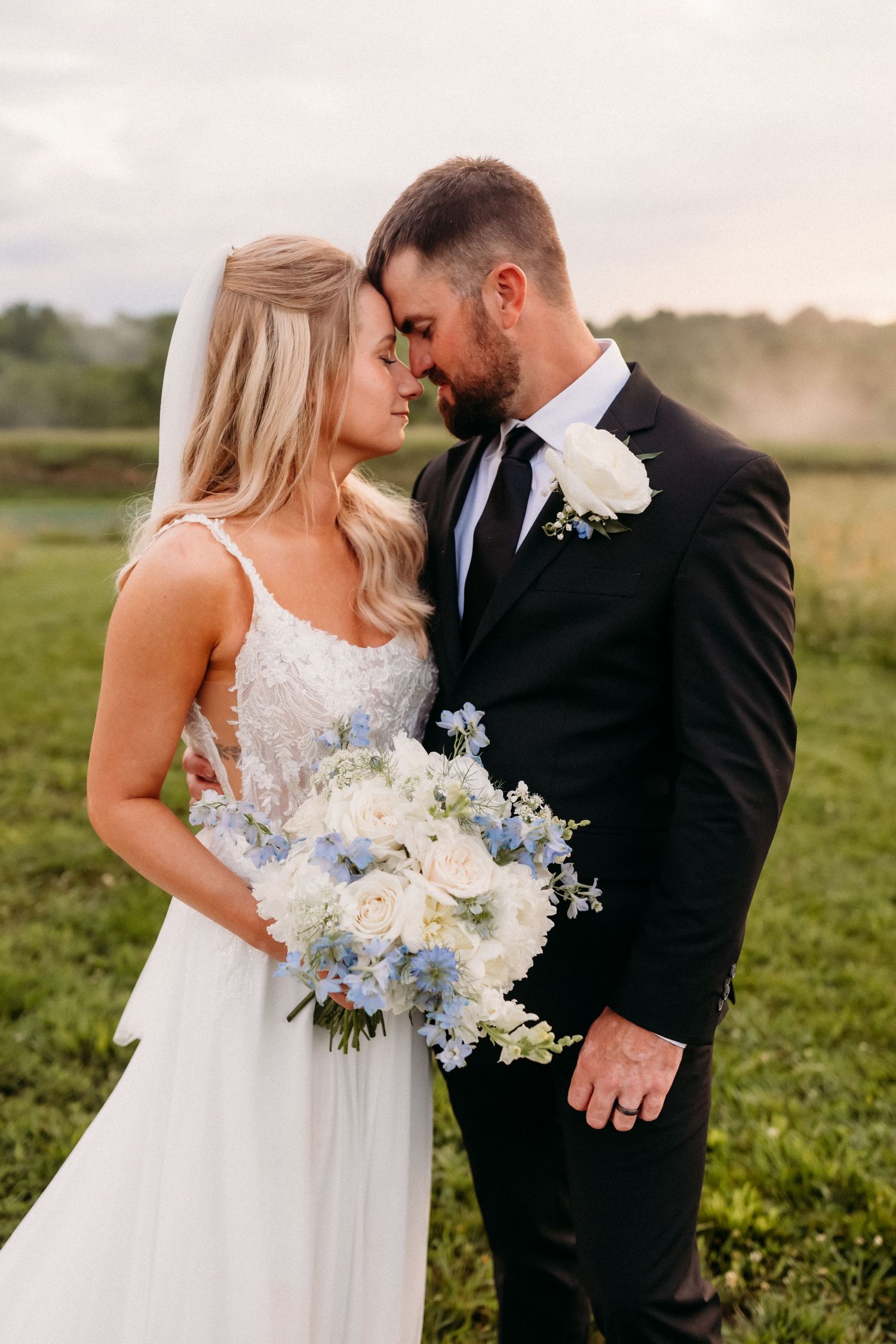 Bride and groom share romantic forehead touch outdoors, bride holding white and blue floral bouquet.