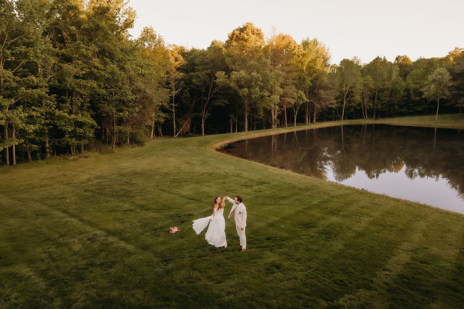Aerial view of bride and groom holding hands on lush green lawn beside a serene pond surrounded by trees at golden hour.