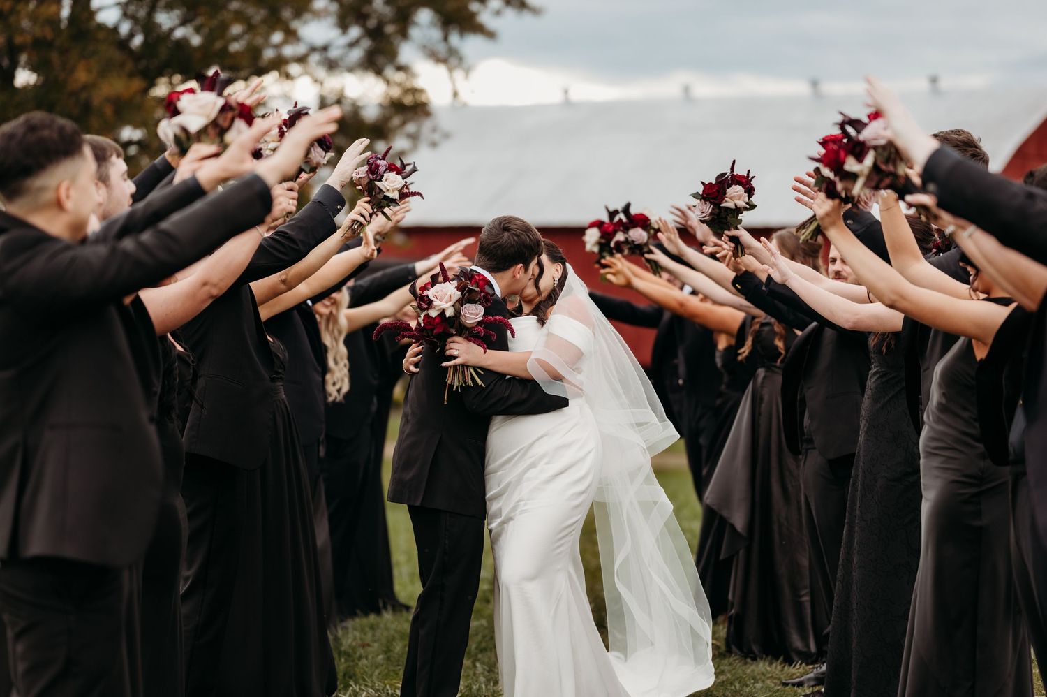 Wedding party in black attire forms arch with bouquets as bride and groom kiss beneath during outdoor ceremony.