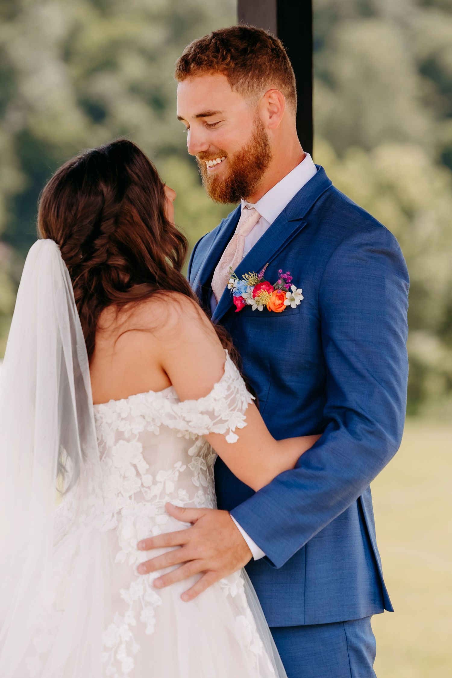 Smiling groom in blue suit with colorful boutonniere embraces bride in off-shoulder lace gown.