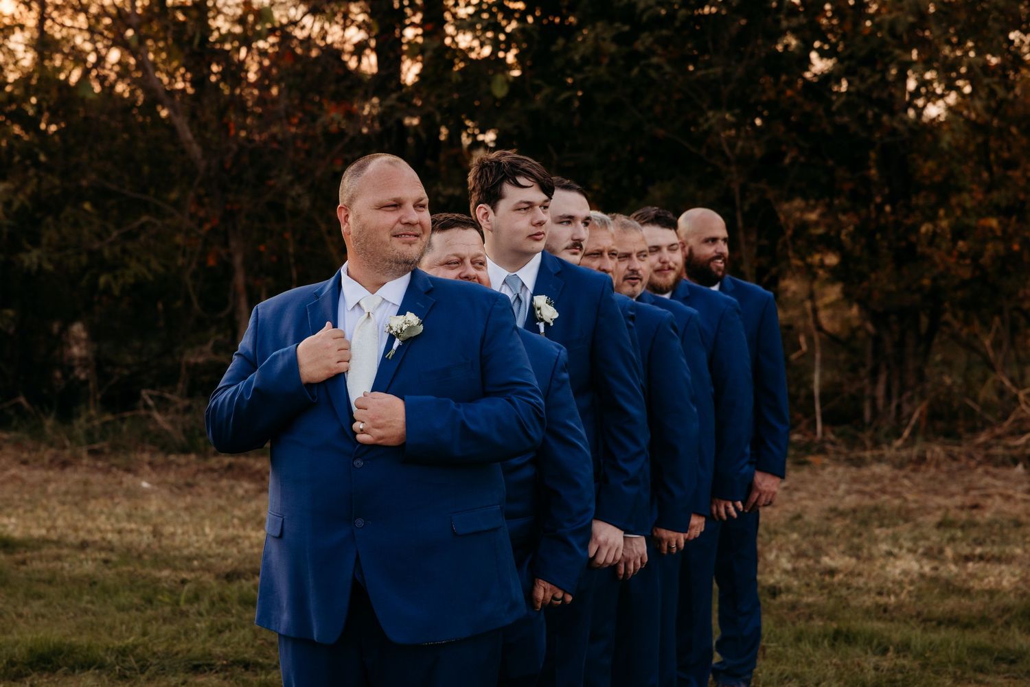 Groom in navy blue suit leads groomsmen in matching suits outdoors at golden hour wedding portrait.