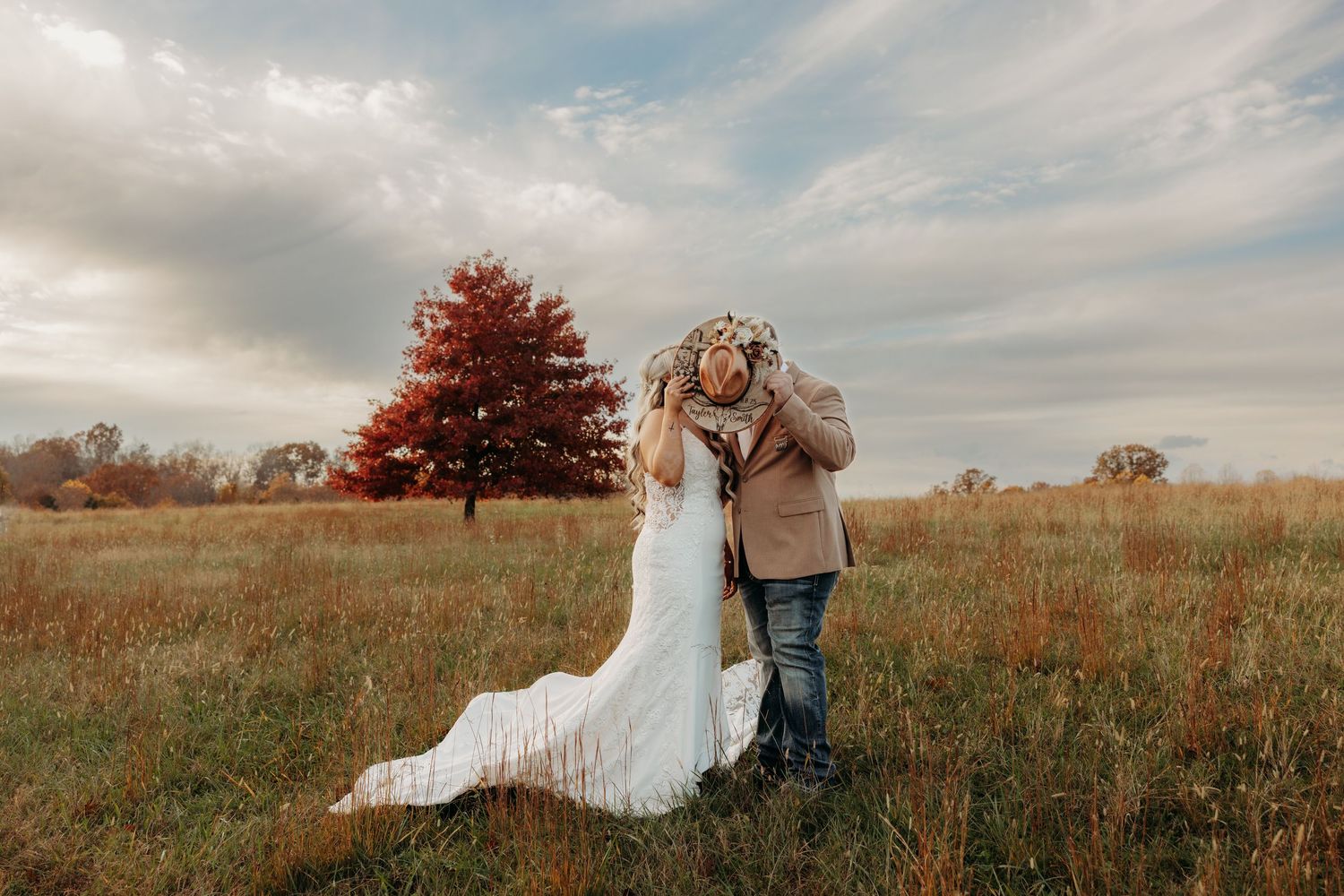 Bride and groom share a romantic moment in an autumn field with a vibrant red tree under a cloudy sky.