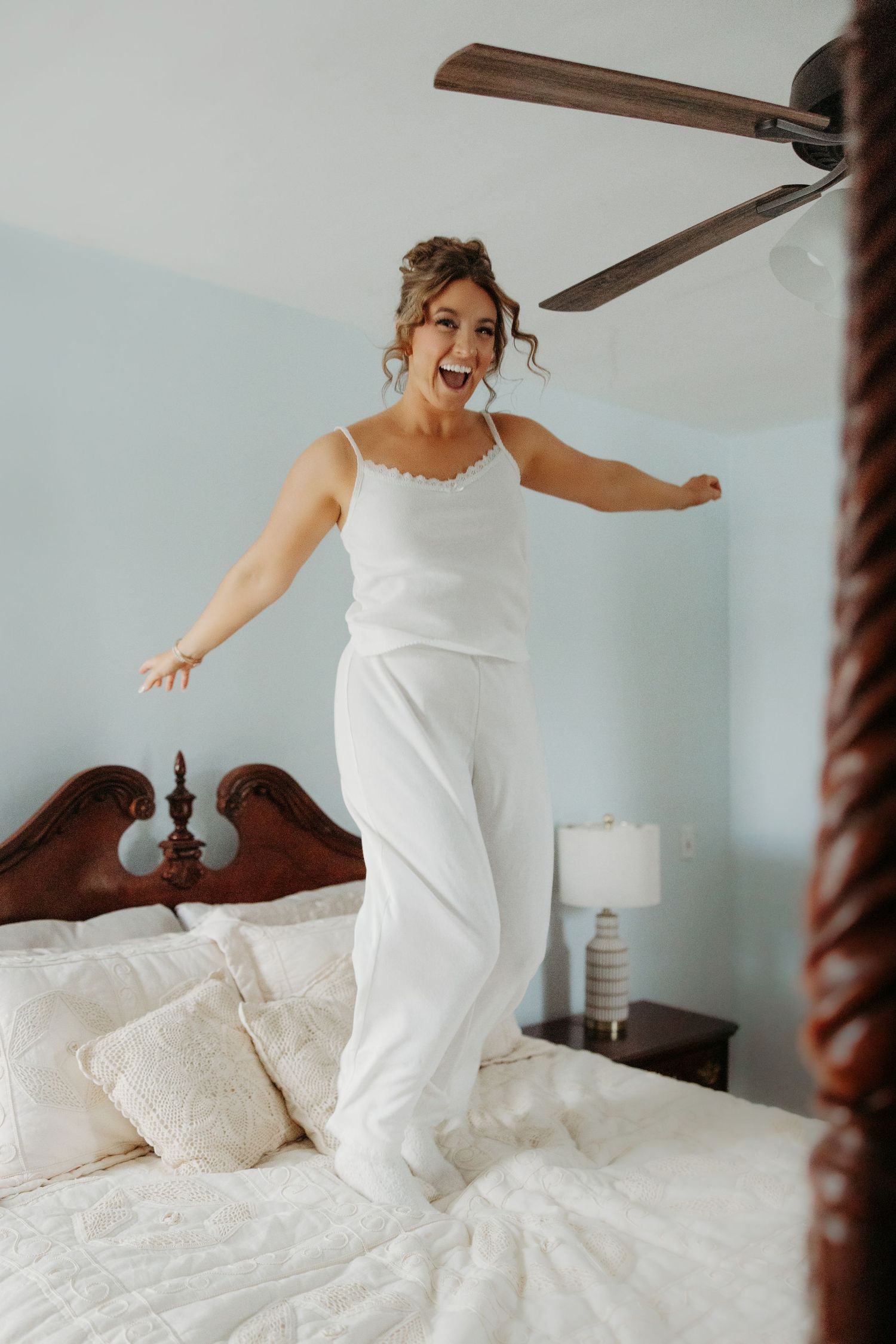 Bride in white pajamas jumping joyfully on bed during morning preparations for wedding day.
