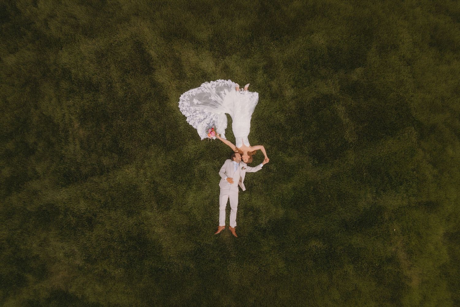 Aerial drone view of bride and groom lying on lush green grass, wedding couple holding hands from above.