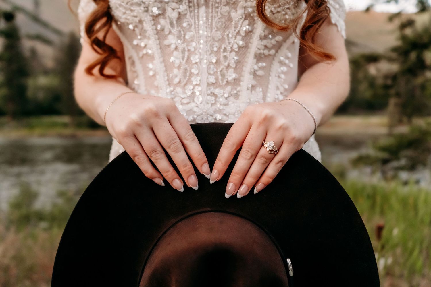 Bride in beaded corset gown holds black hat, showcasing elegant ring and bracelet with soft pink manicure.