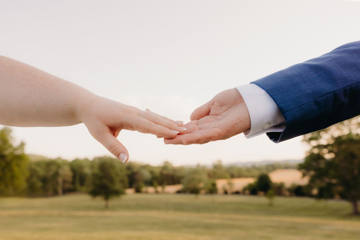 Bride and groom gently touching fingertips outdoors with a lush green park landscape in the background.