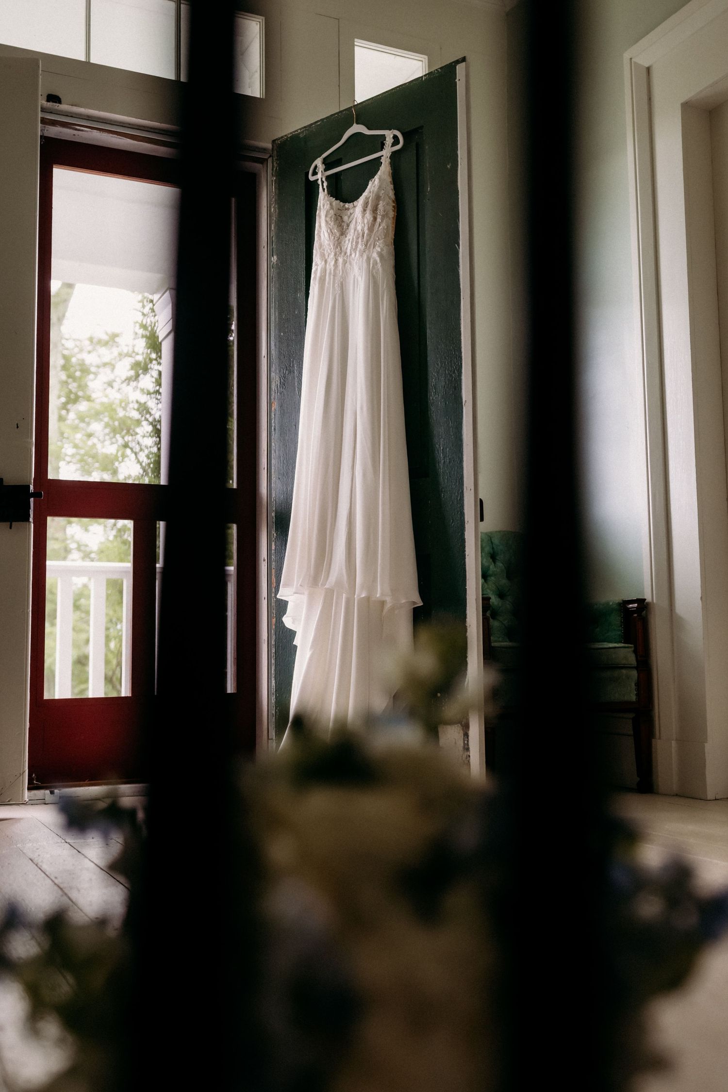 White lace wedding dress hanging on a green door in a rustic venue with natural light.