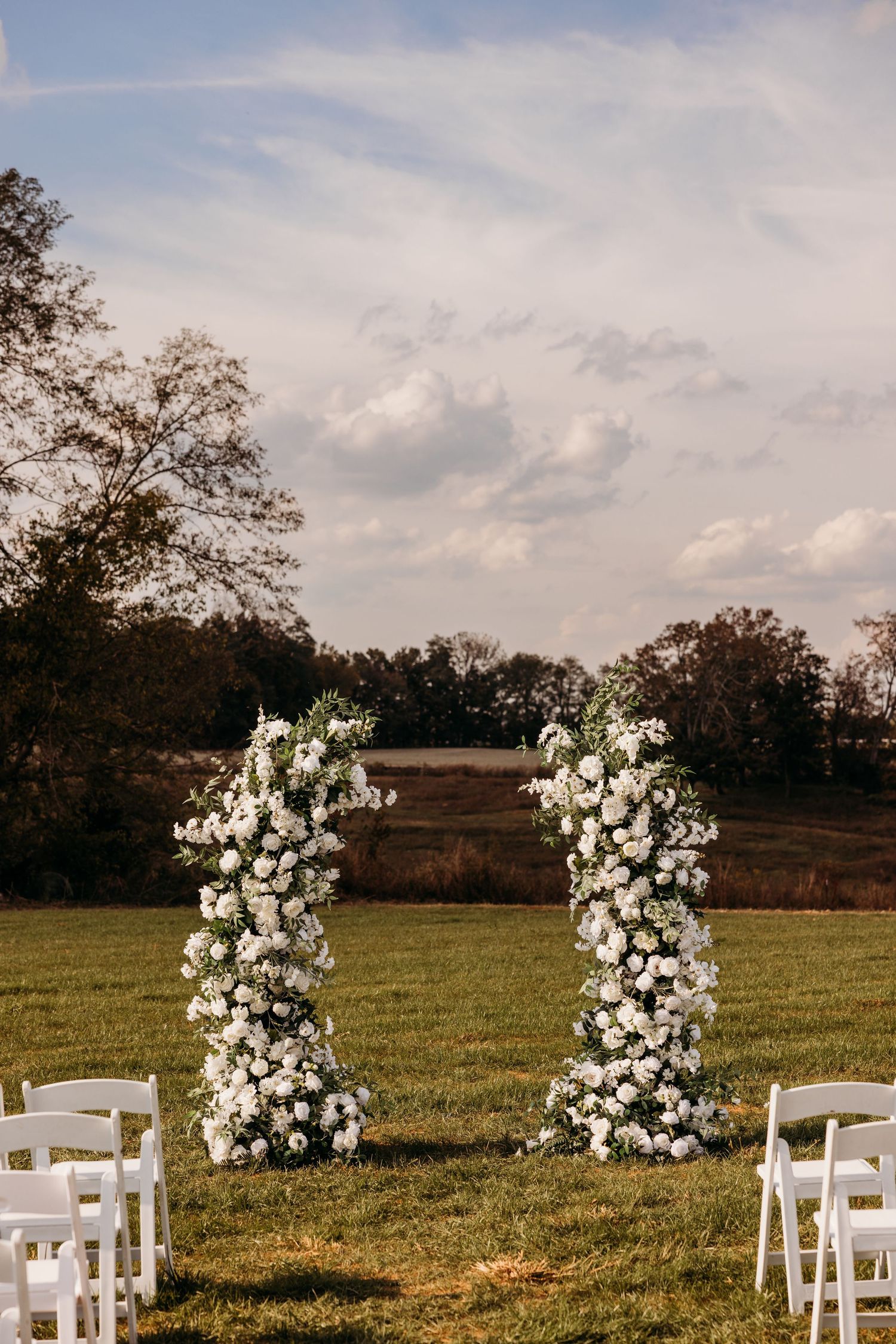 Two white floral columns adorned with roses create an elegant outdoor wedding ceremony arch on a green lawn.