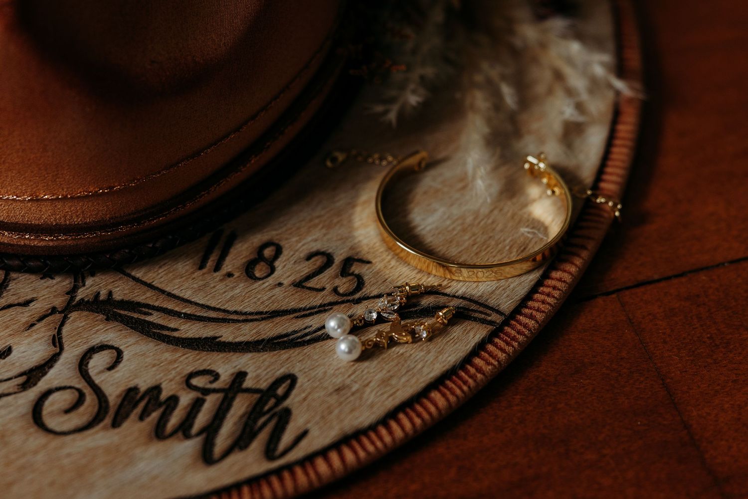 Gold wedding ring and pearl earrings rest on engraved wooden keepsake with Smith name, date 11.8.25, beside cowboy hat.