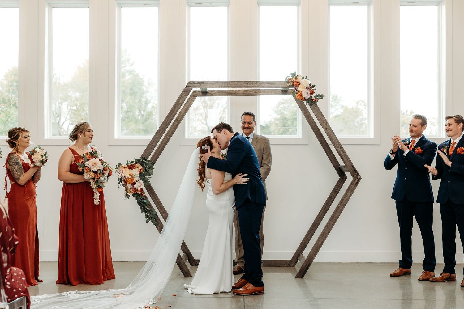 Bride and groom share first kiss under hexagonal wooden arch at bright white wedding venue ceremony.