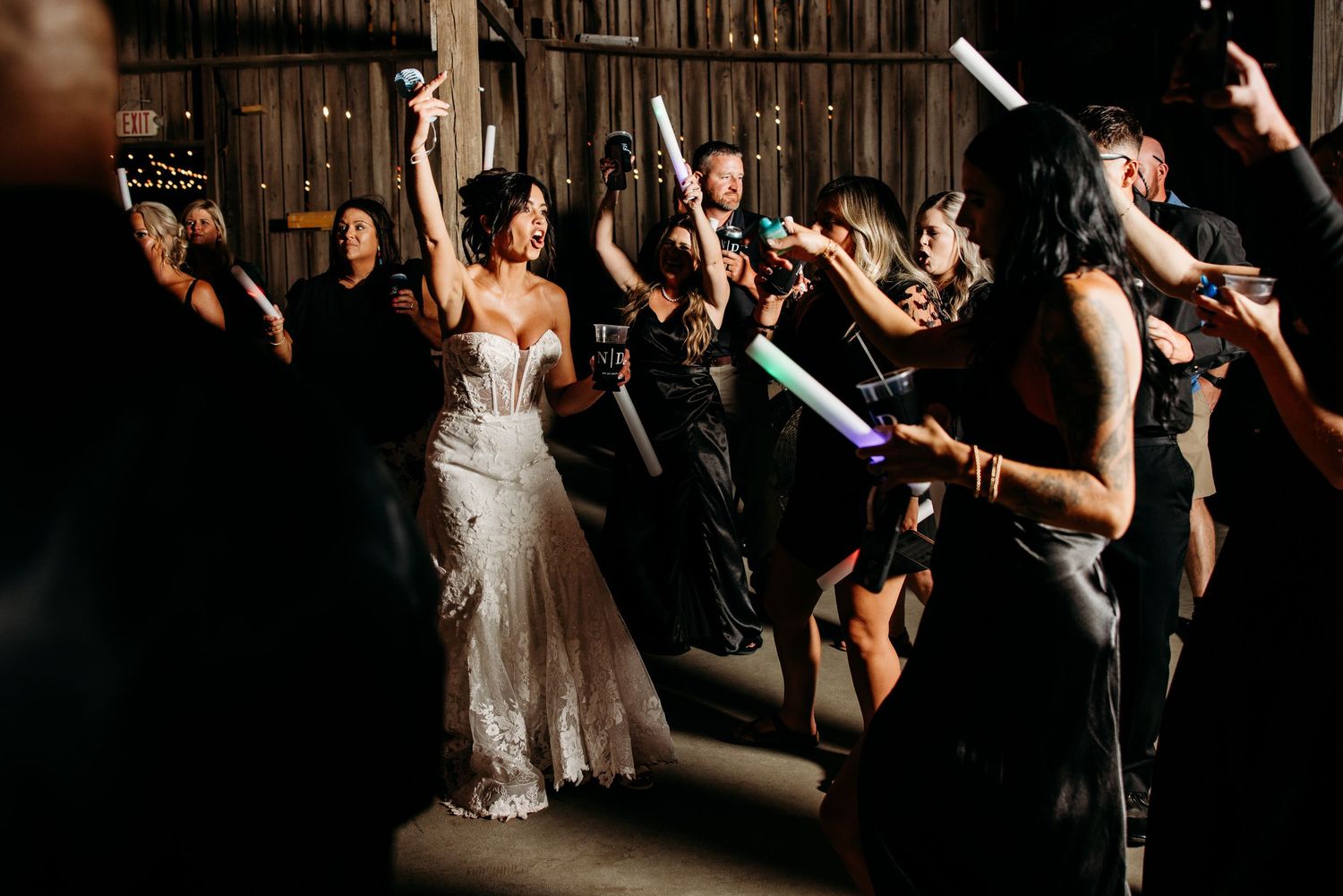 Bride celebrates with guests waving glow sticks during a lively wedding reception in a rustic barn.