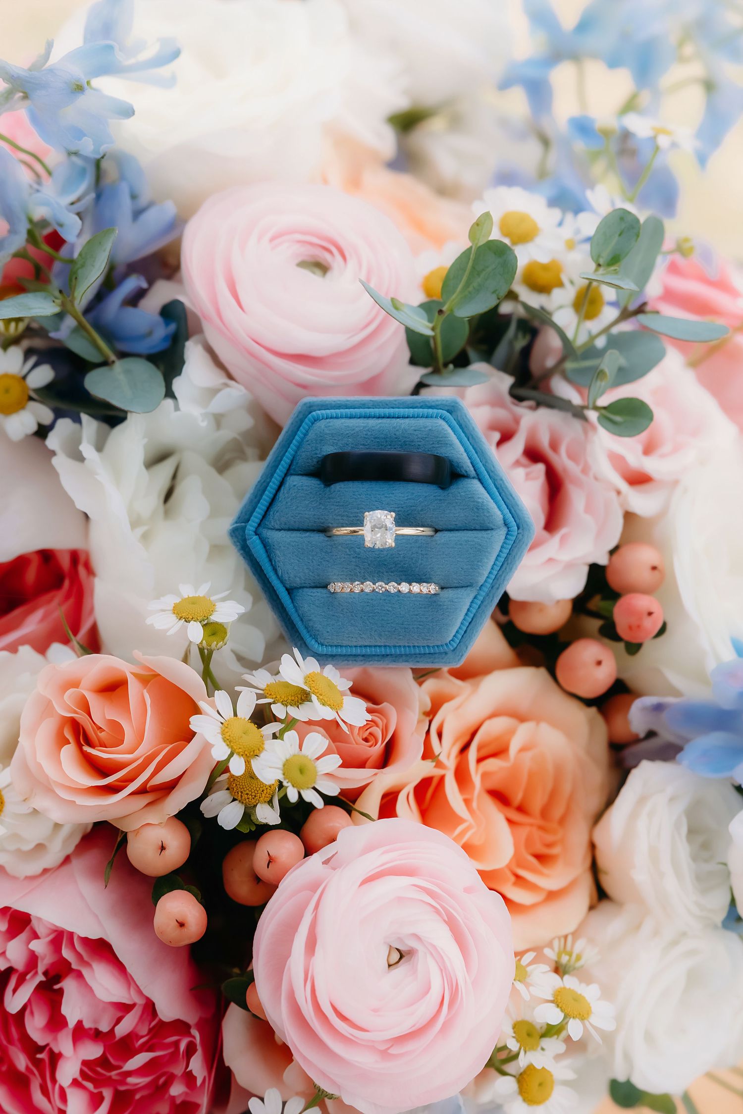 Blue velvet ring box with diamond engagement ring and wedding band resting on a colorful bridal bouquet of pink roses.