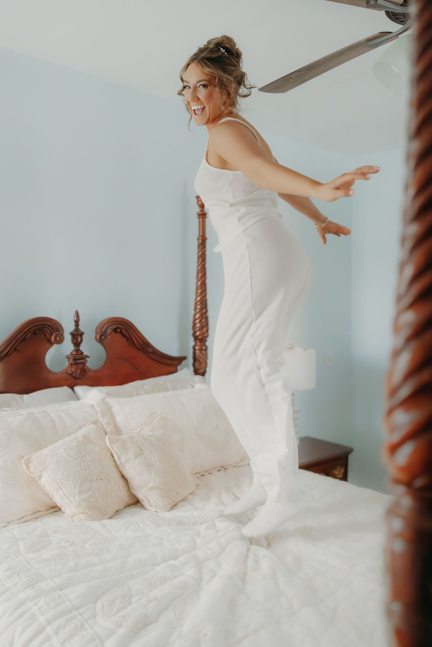 Joyful bride in white gown dancing on bed during wedding morning preparations.