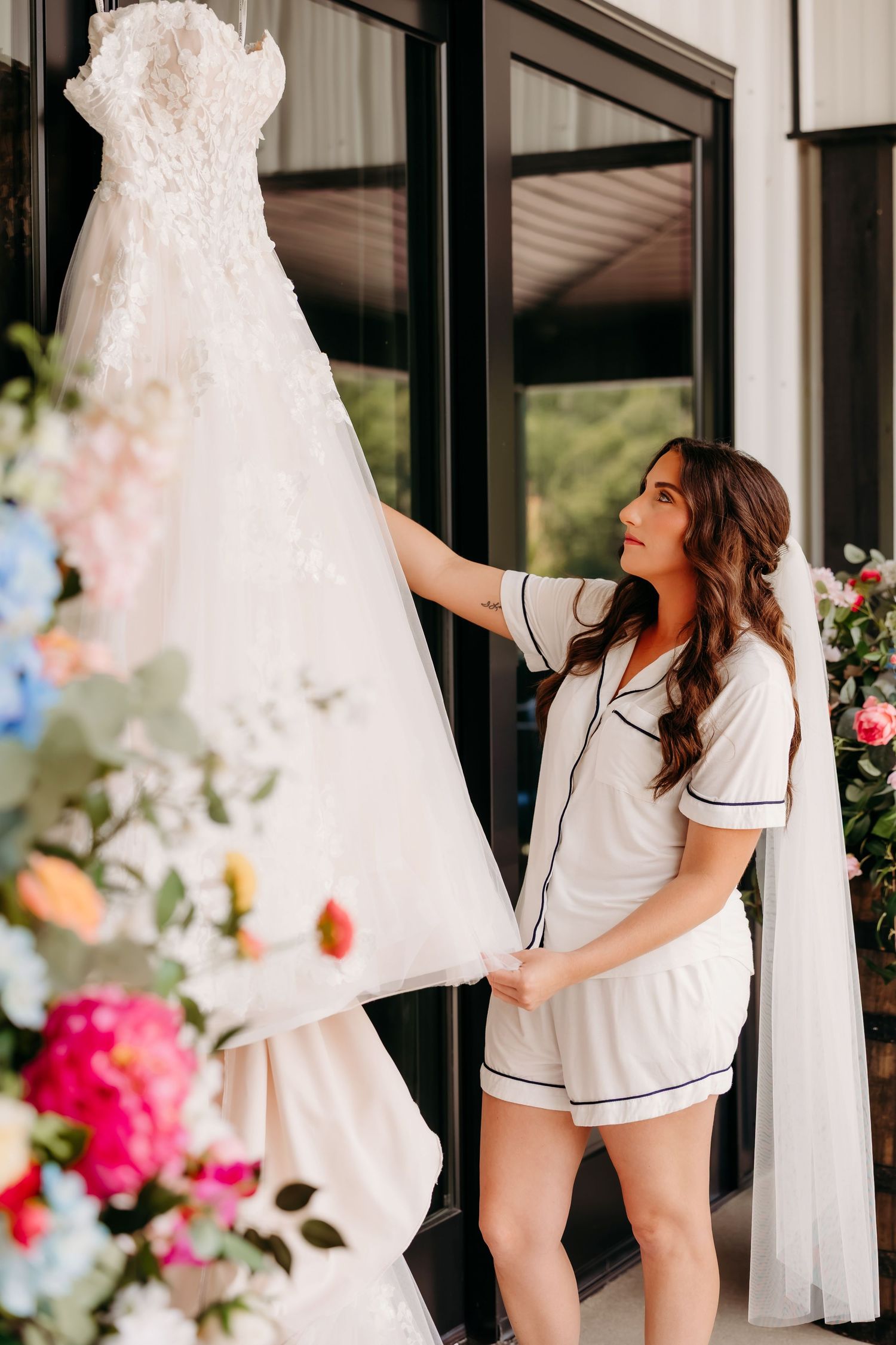 Bride in white pajamas admiring her lace wedding dress hanging near a window with colorful flowers nearby.