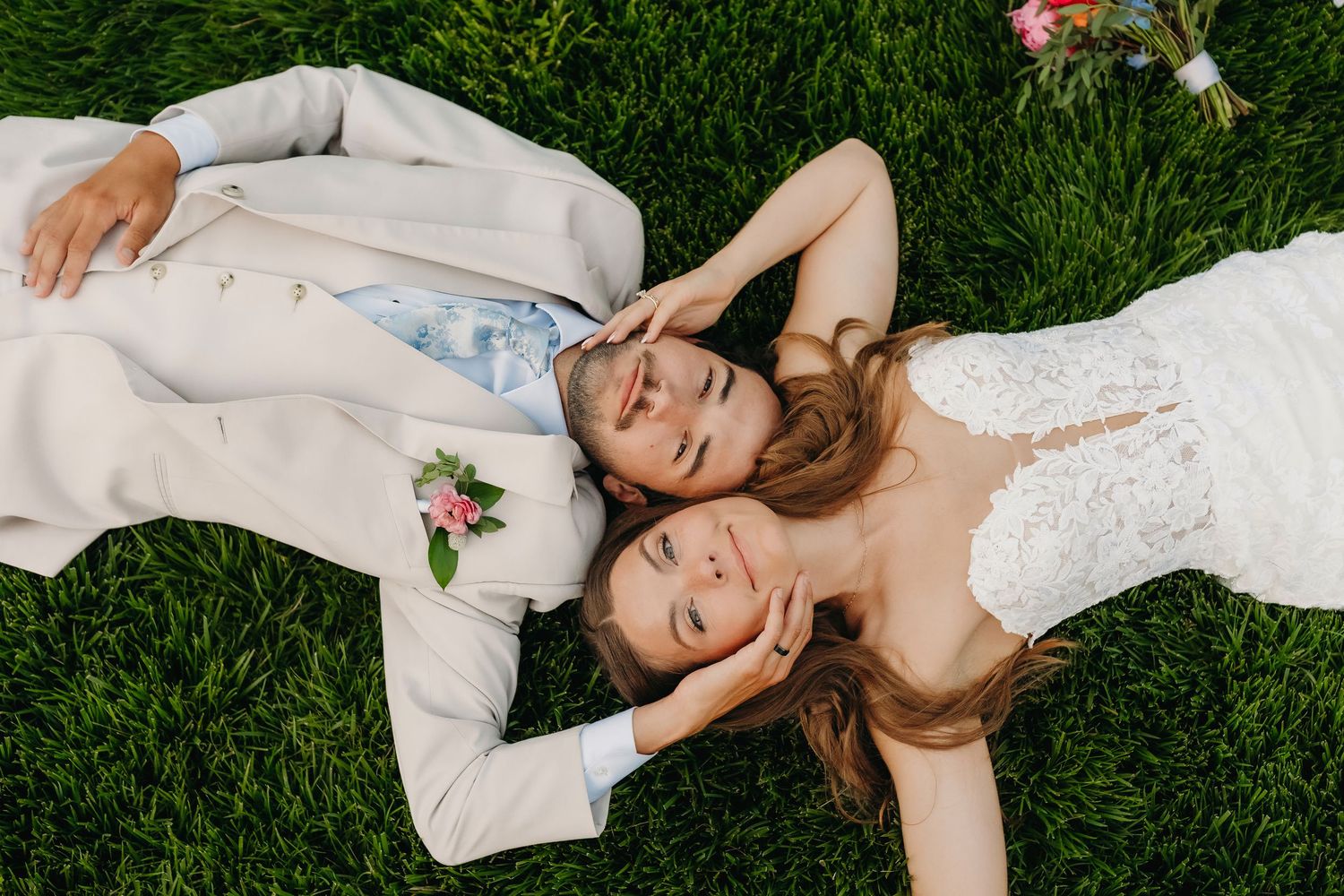 Bride and groom lying on green grass, heads together, smiling, wearing white wedding attire with floral boutonniere.