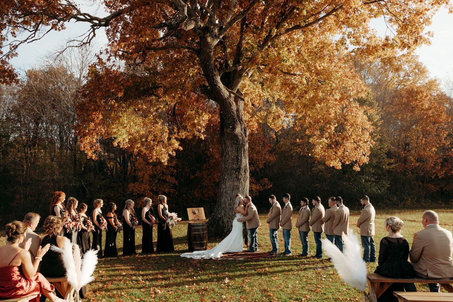 Outdoor fall wedding ceremony under a large oak tree with golden leaves, bridal party lining the aisle.