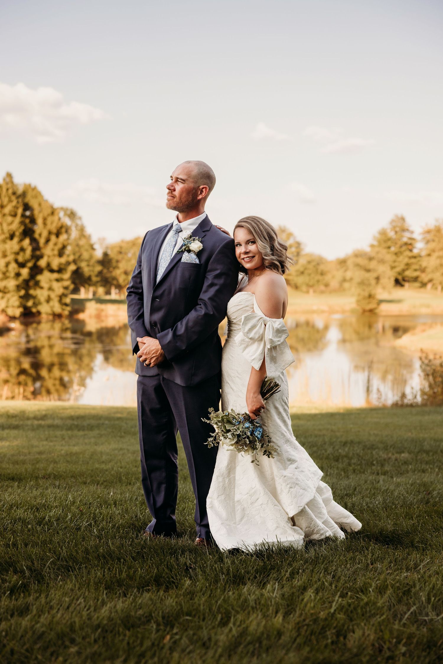 A romantic sunset wedding photo of a couple walking together on a grassy field near a lake.