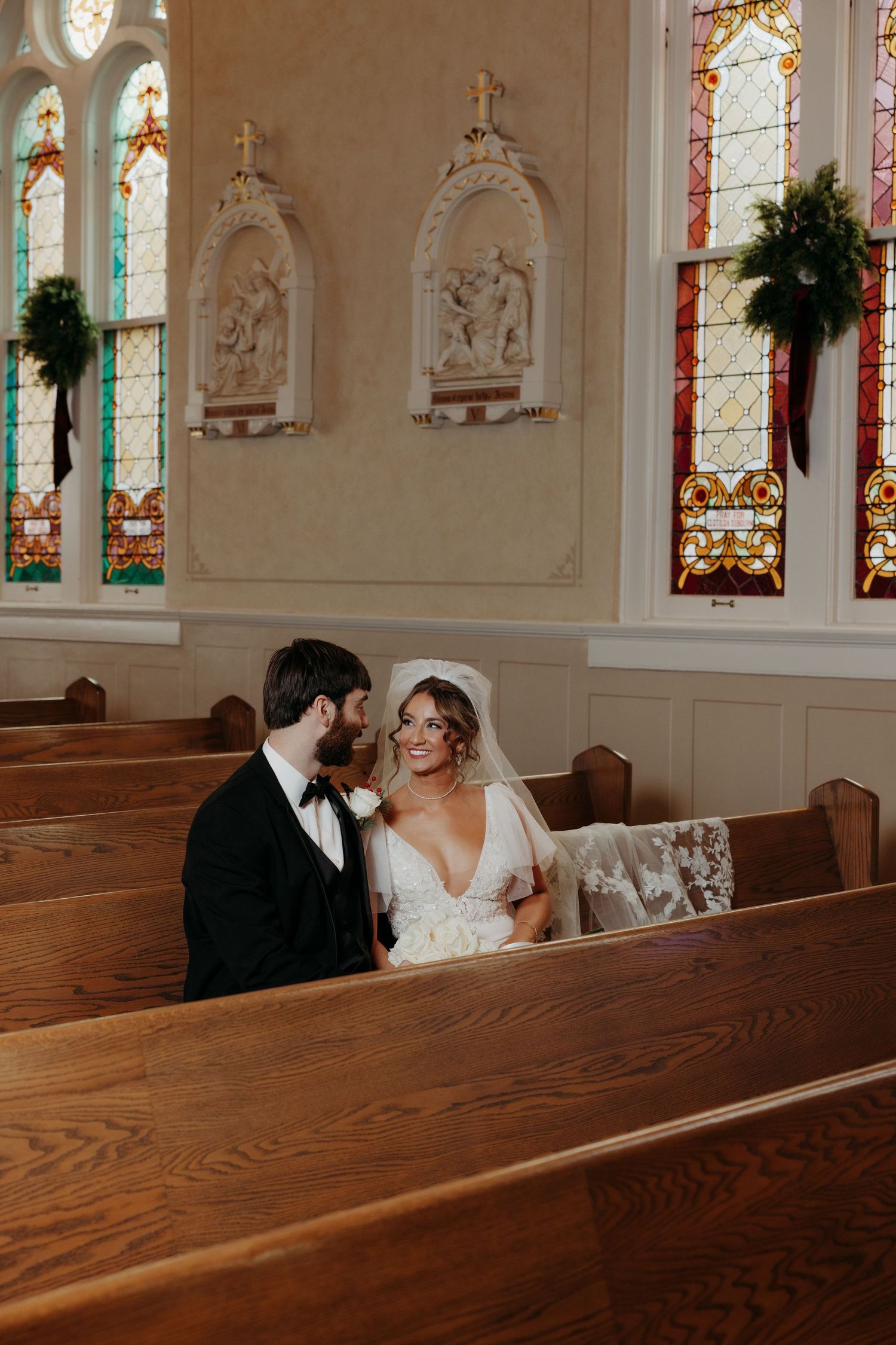 A romantic wedding moment captured in wooden church pews with stained glass windows in the background.