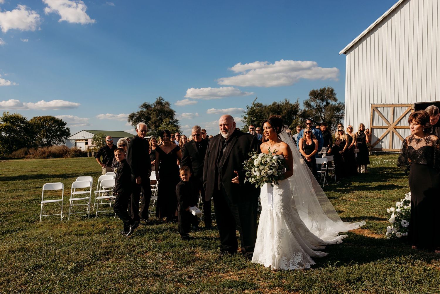 Bride walks down outdoor aisle with escort at rustic barn wedding ceremony on sunny day.