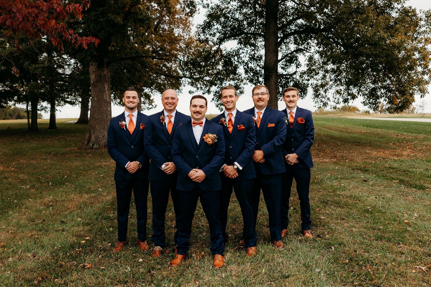 Groom and groomsmen in matching navy suits with orange bow ties pose outdoors on autumn wedding day.