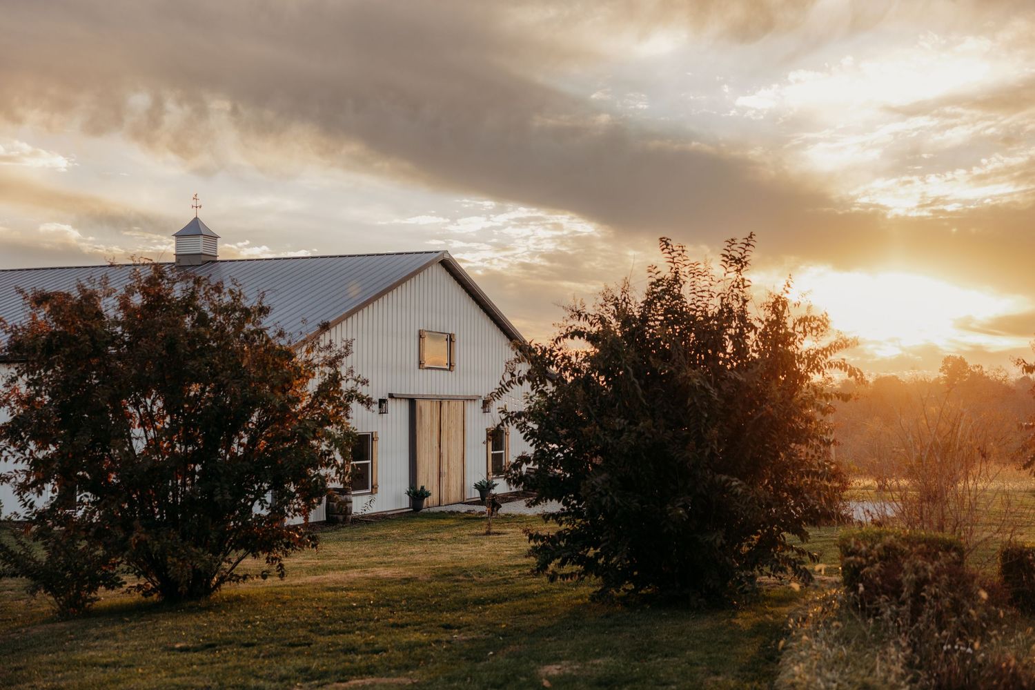White barn wedding venue glowing at golden sunset with dramatic clouds and lush green trees.