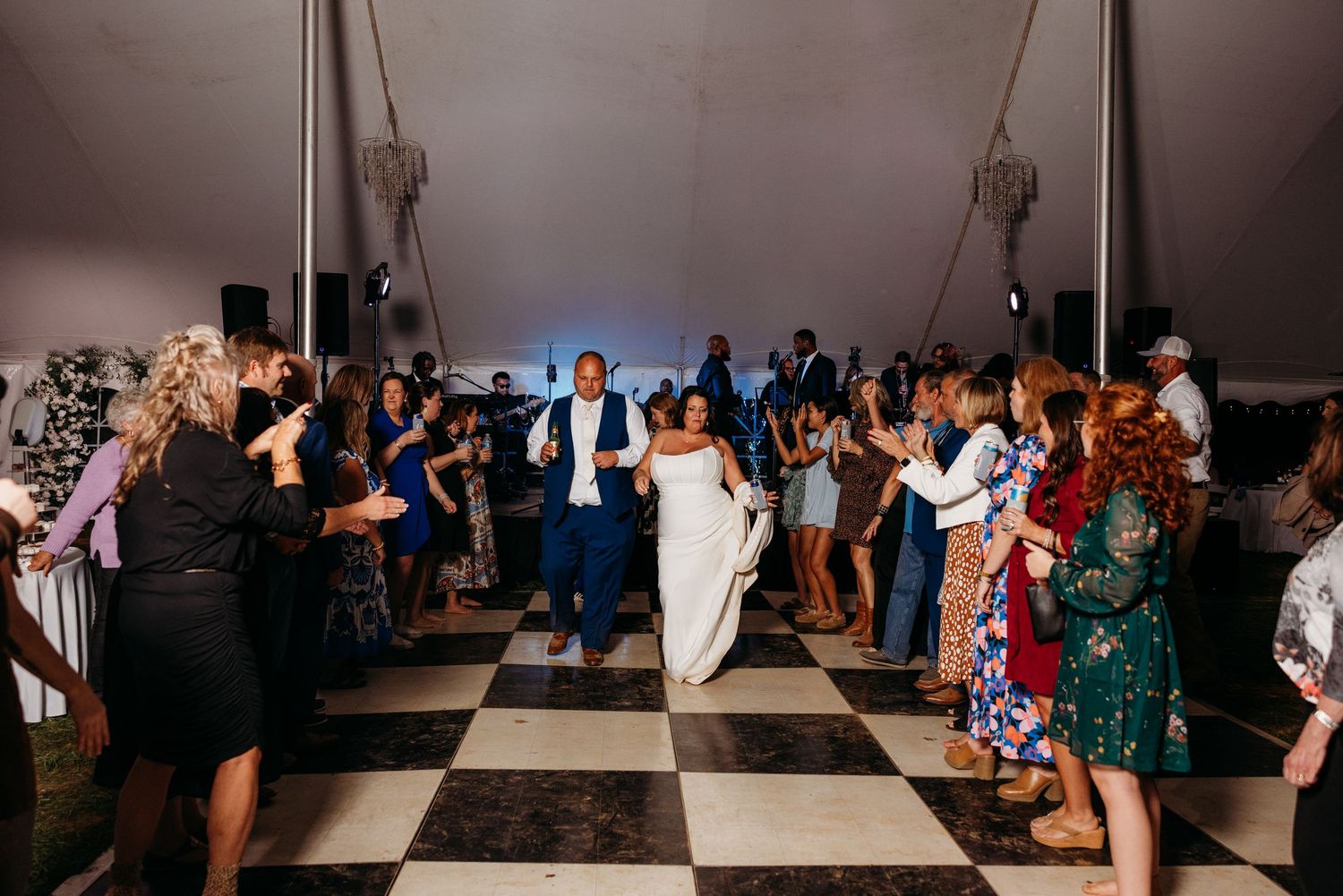 Bride and groom enter wedding reception on checkered dance floor surrounded by cheering guests under a white tent.