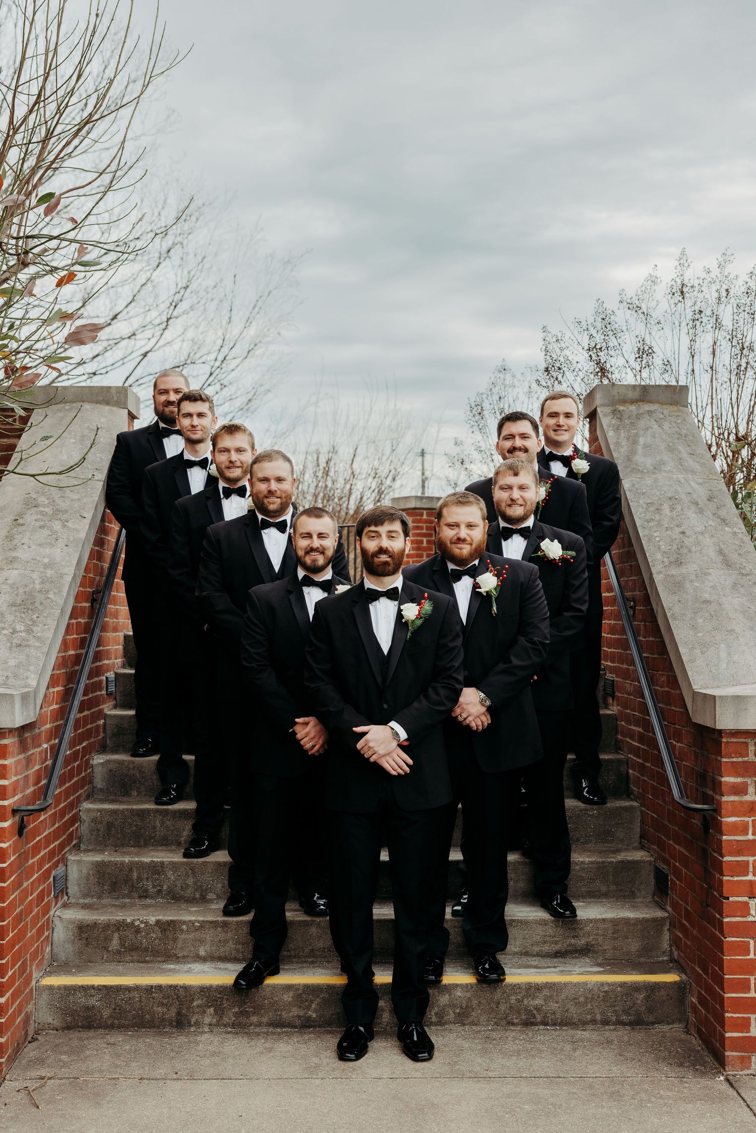 Groom and groomsmen in black tuxedos with boutonnieres pose on outdoor stone steps for a wedding photo.