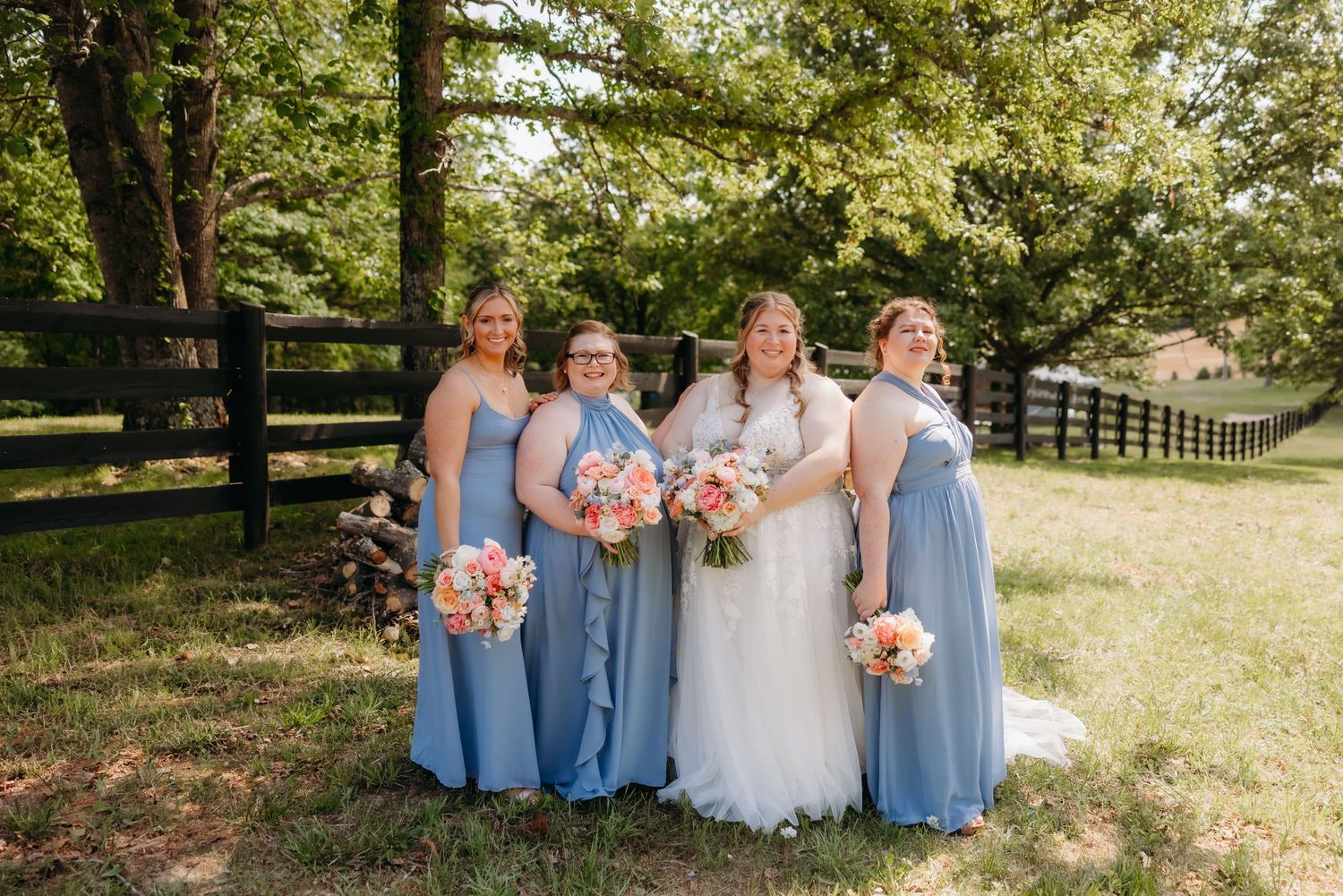 Bride with three bridesmaids in blue dresses holding pink bouquets outdoors at wedding venue.