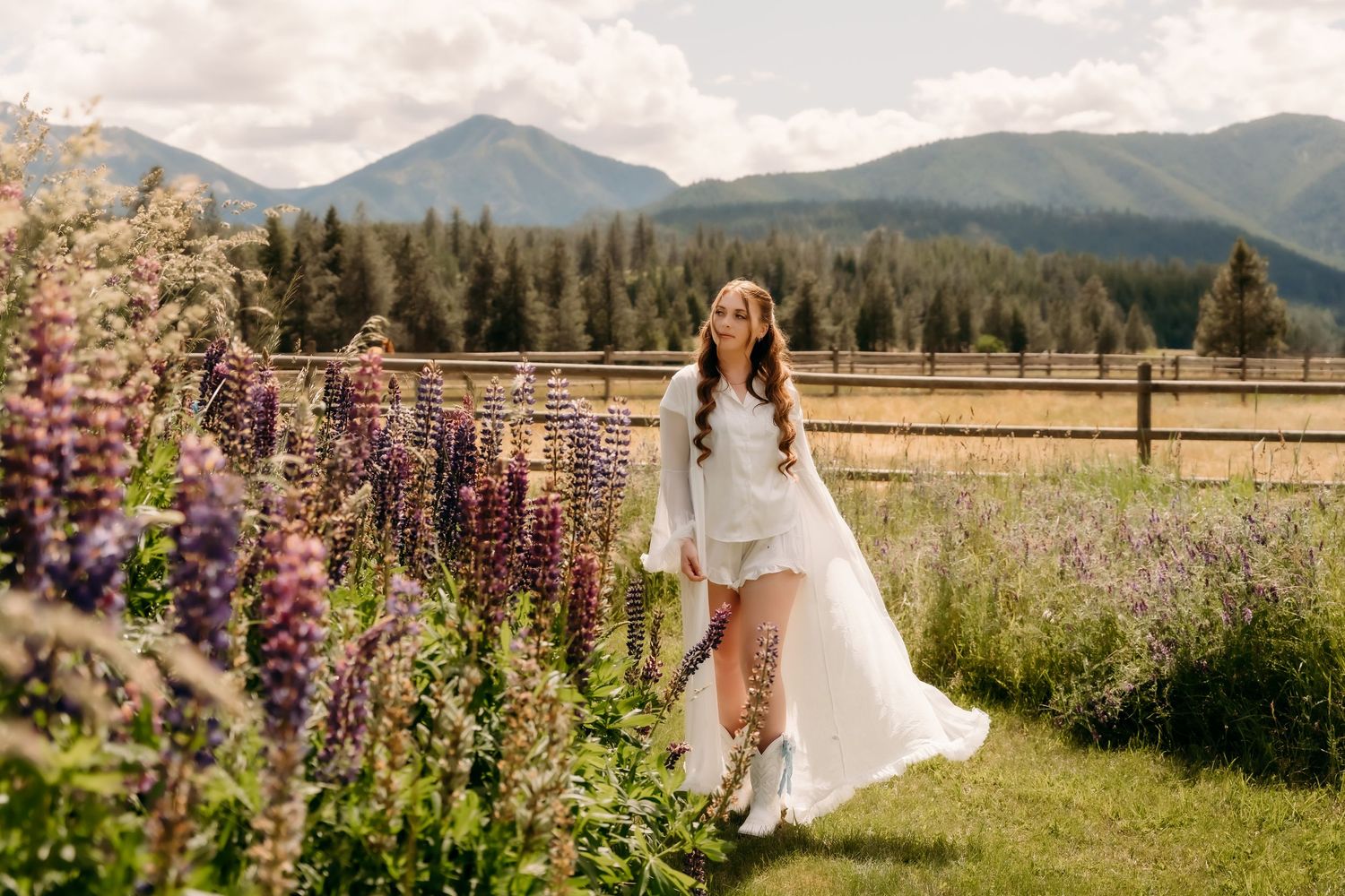 Bride in white dress walks among colorful lupine flowers with mountain backdrop on sunny day.