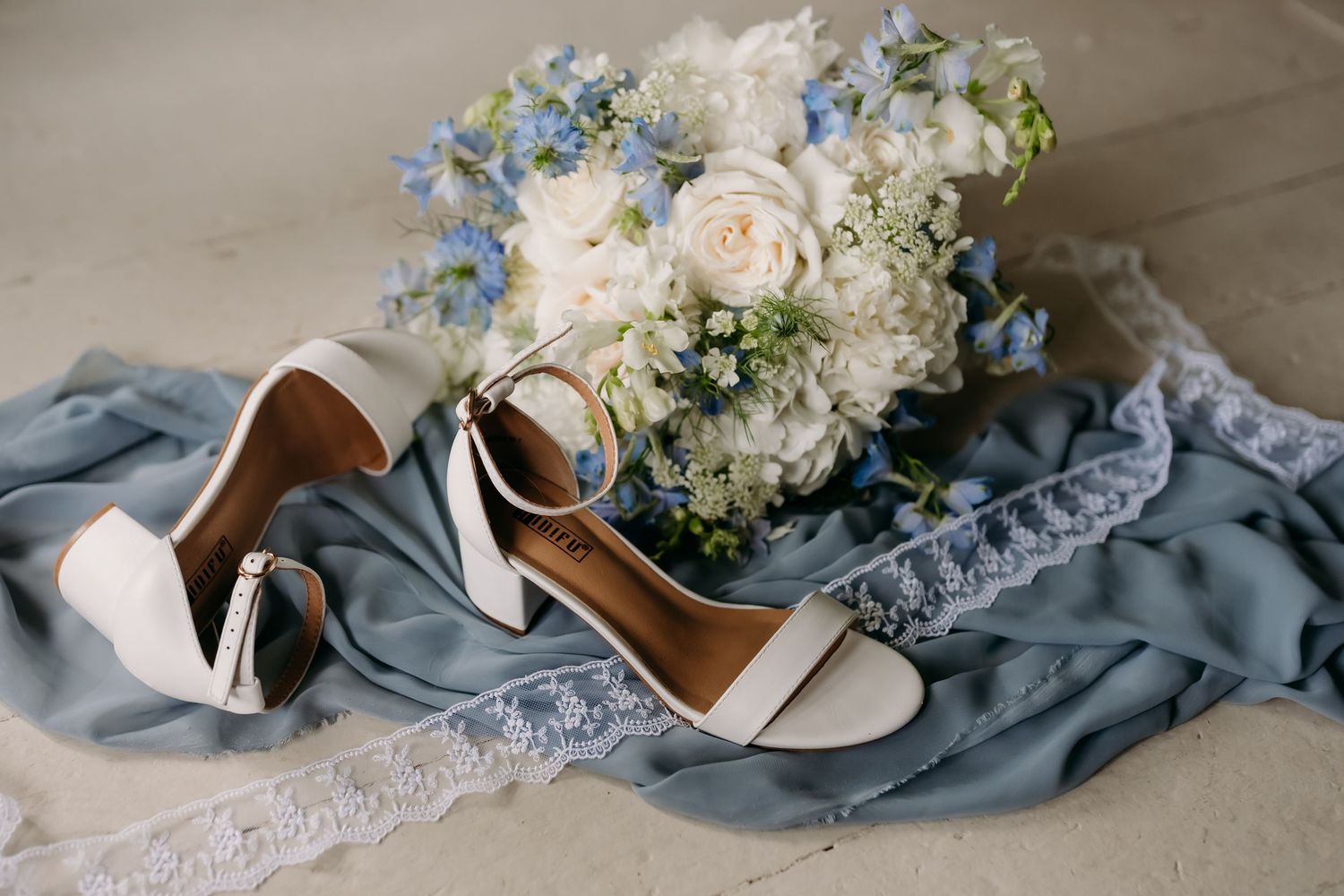 White bridal heels beside a blue and white floral bouquet on dusty blue chiffon fabric with lace trim.