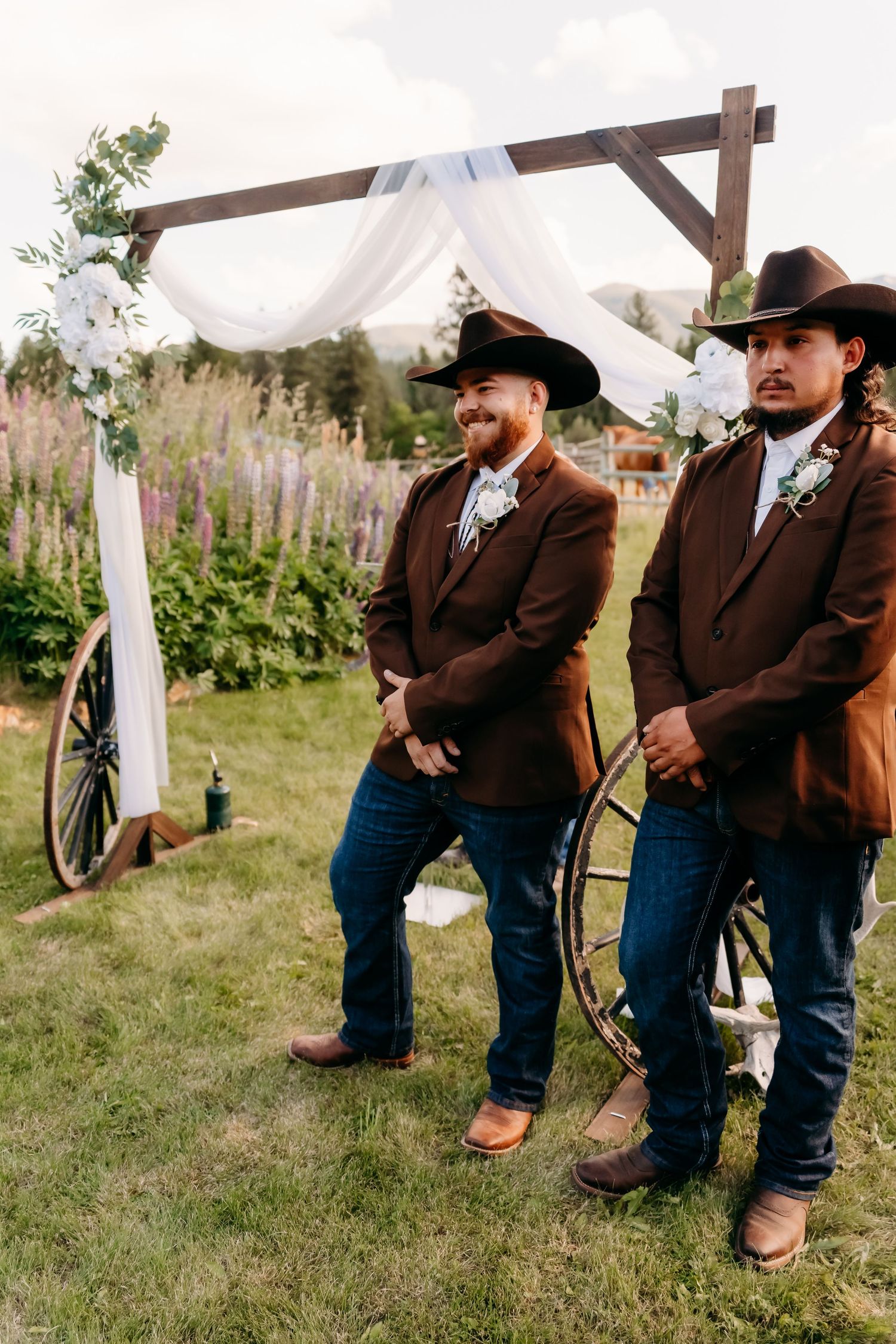 Two grooms in brown suits and cowboy hats stand at outdoor rustic wedding altar with white draping and floral decor.